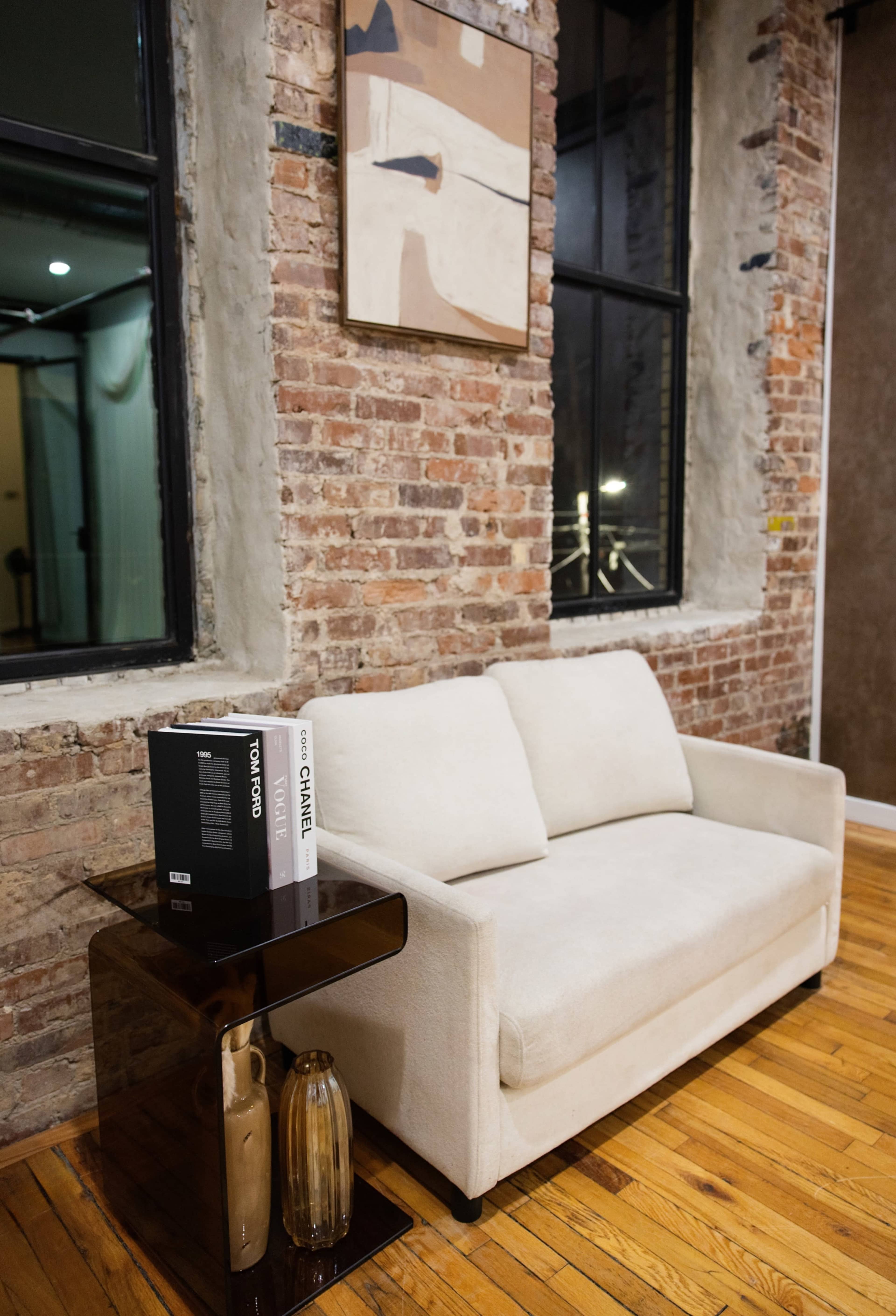 A light-colored sofa is positioned against a brick wall, with a nearby side table holding a black book and a decorative vase.