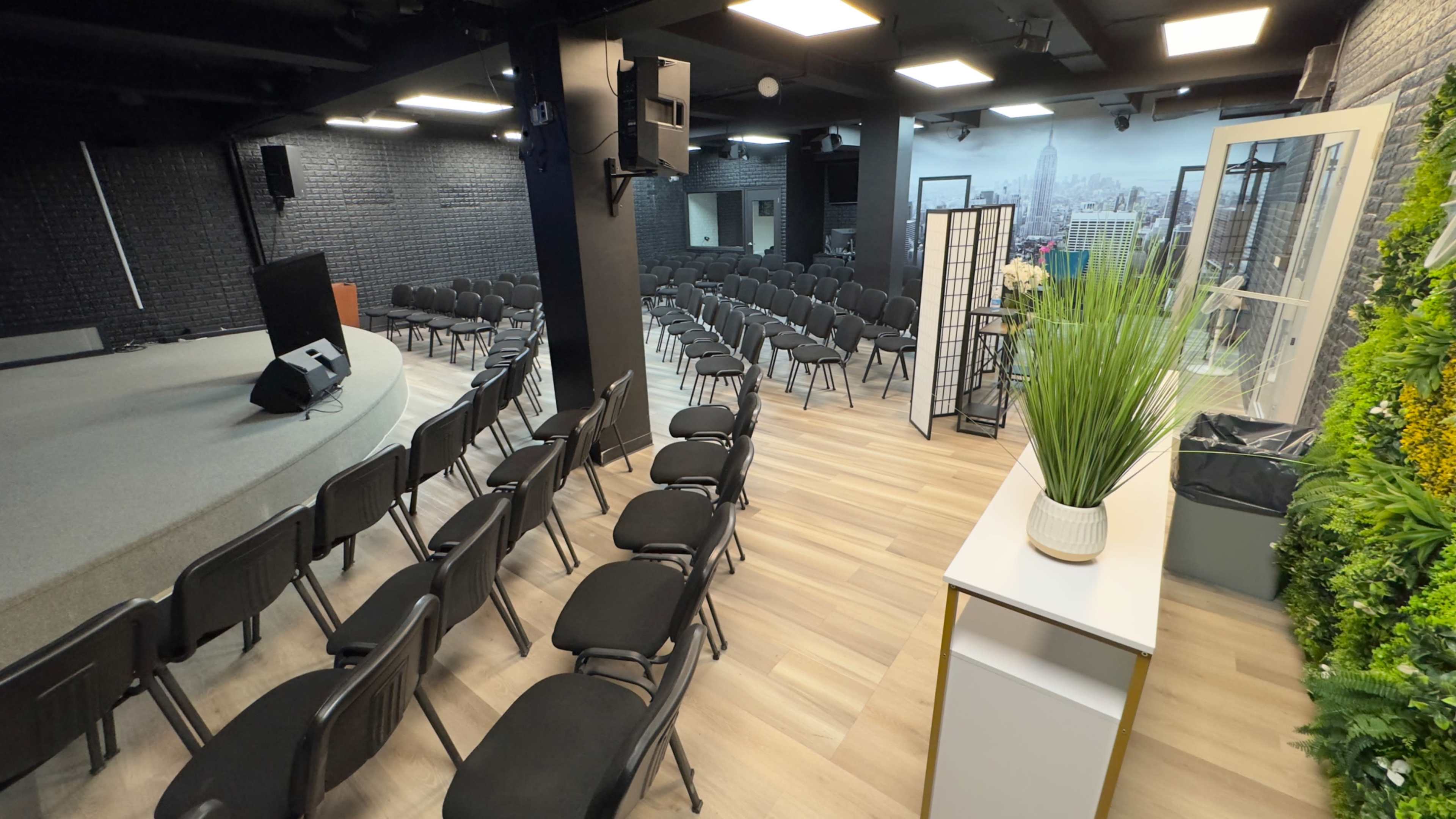 A spacious indoor venue set up with rows of black chairs facing a stage area, with a decorative plant and partition in the foreground.