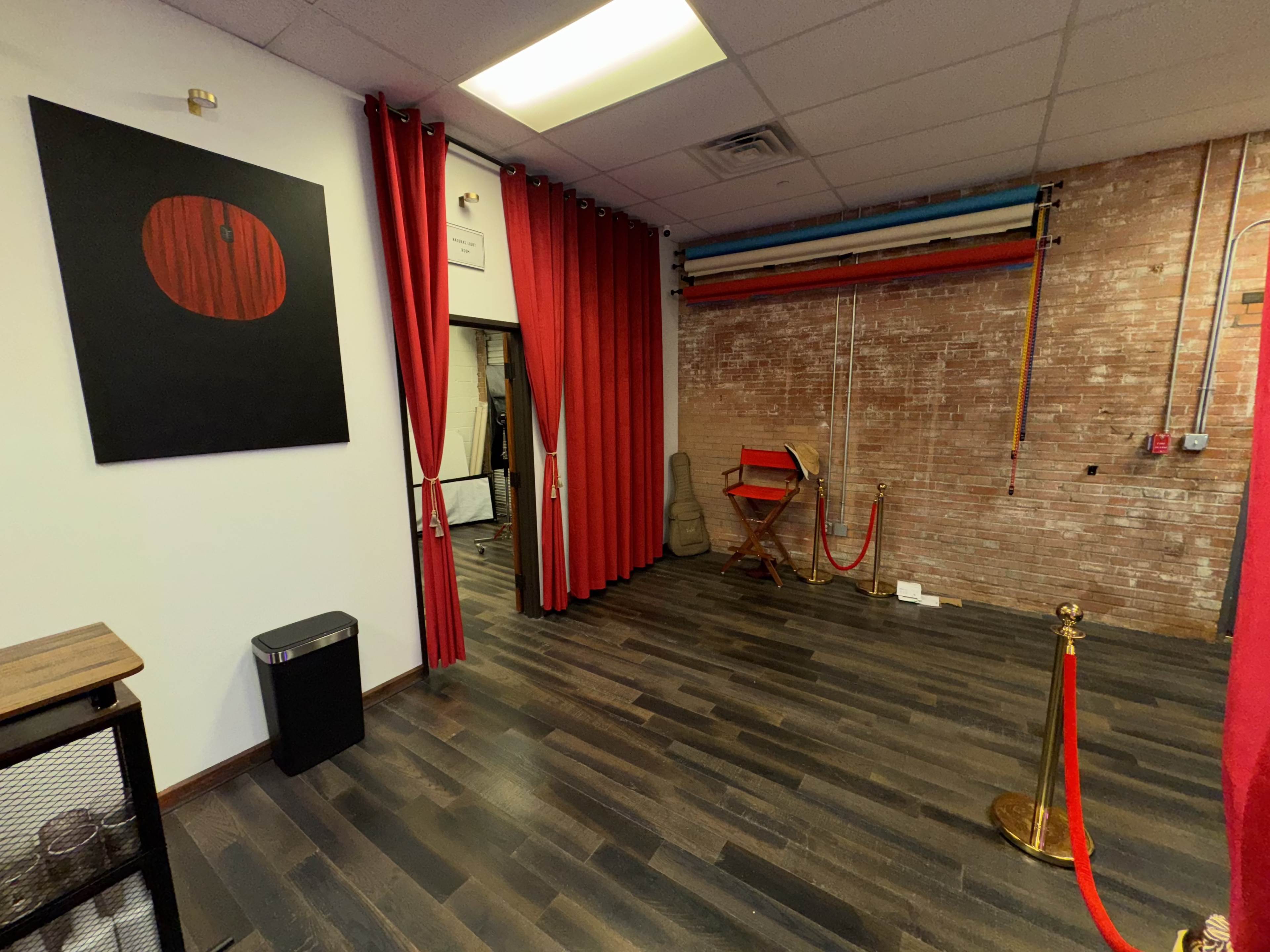 The image shows a room with red curtains, a wooden floor, and a brick wall, featuring a black artwork on the wall and a red director's chair along with a stanchion with a rope.