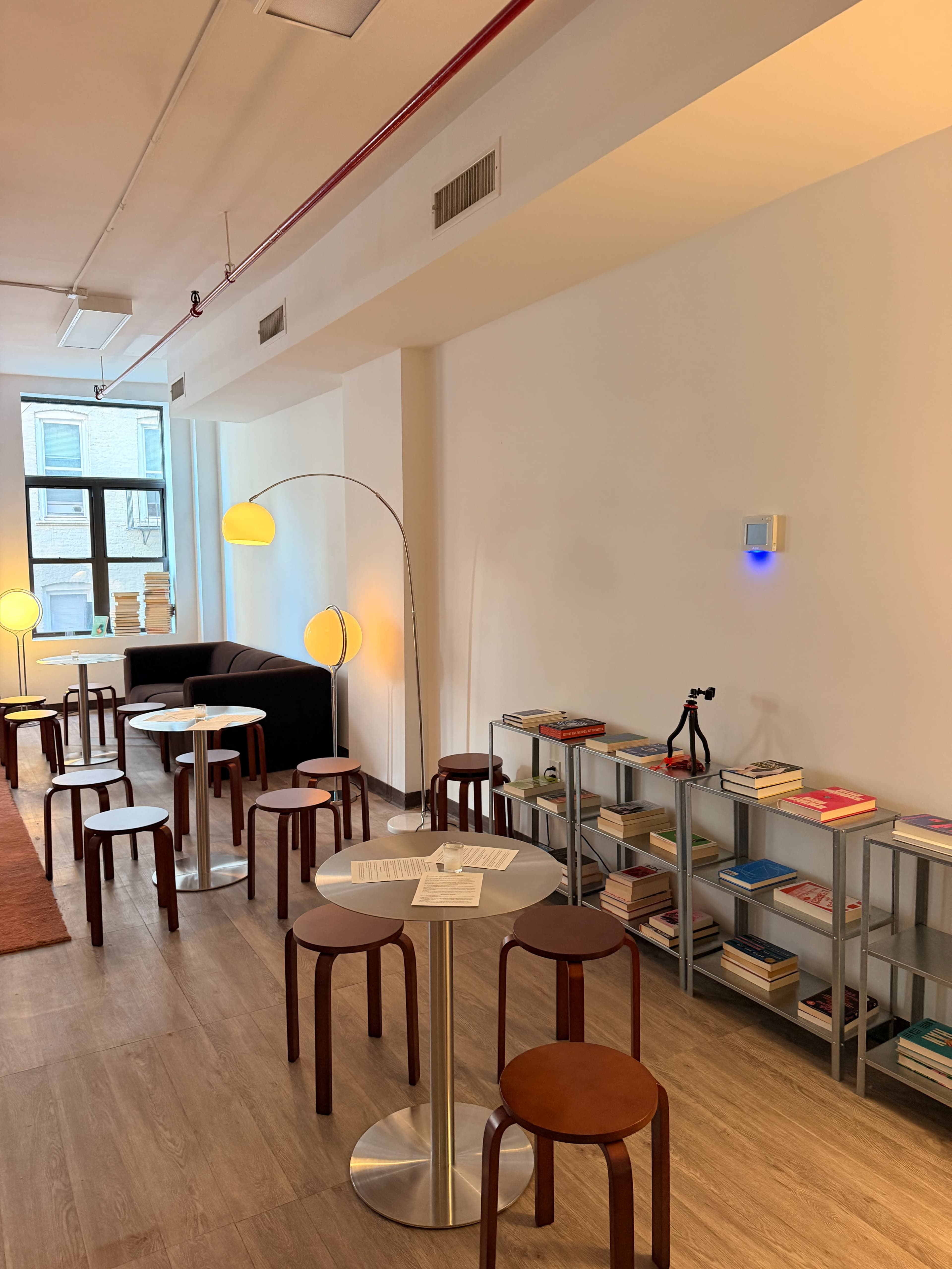 The scene features a minimalist reading area with several small tables and stools, a couch, and a bookshelf containing neatly arranged books, illuminated by floor lamps.
