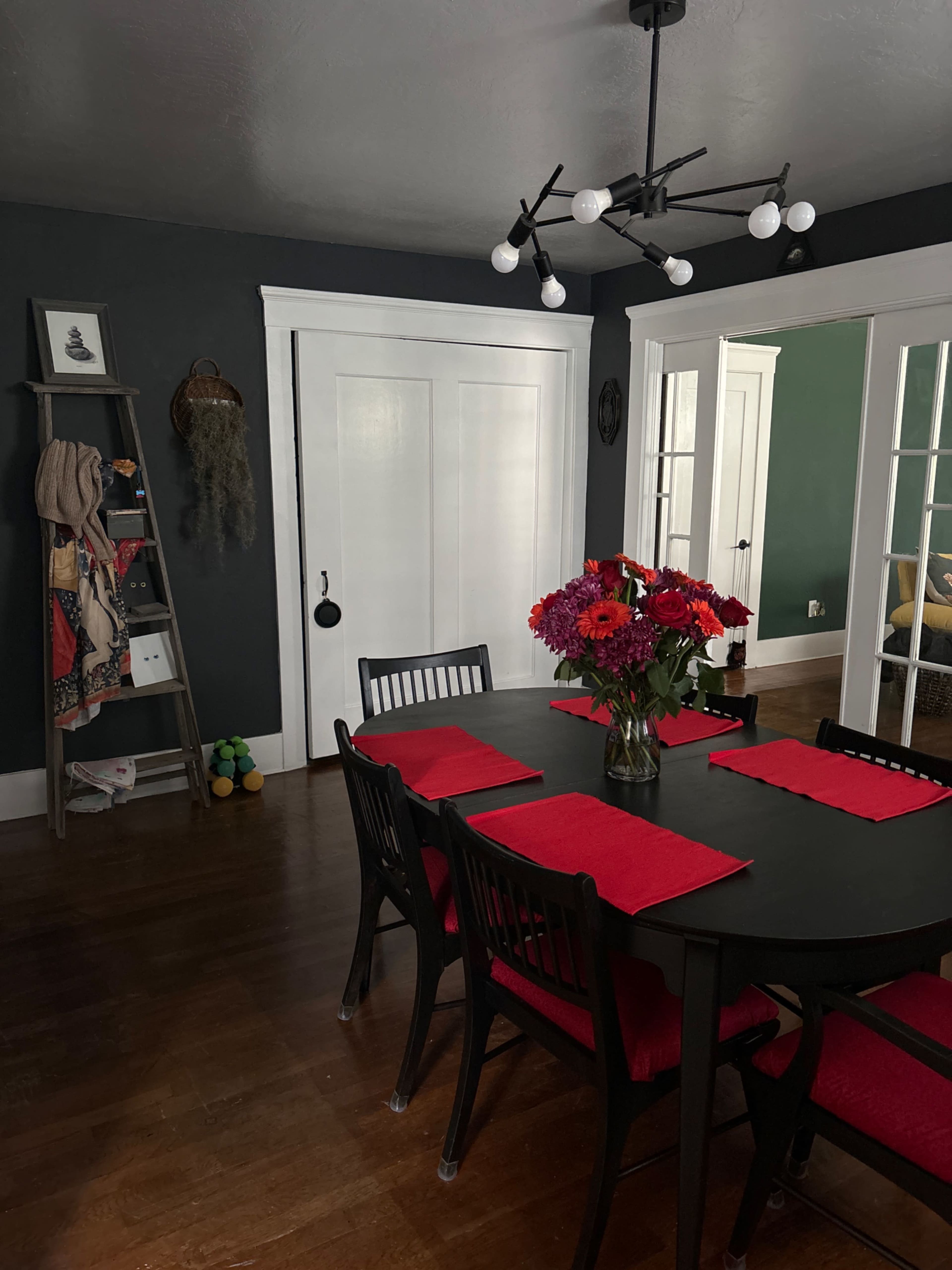 A dining room features a round black table set with red placemats and a floral centerpiece, surrounded by black chairs, with dark walls and a bookshelf in the corner.