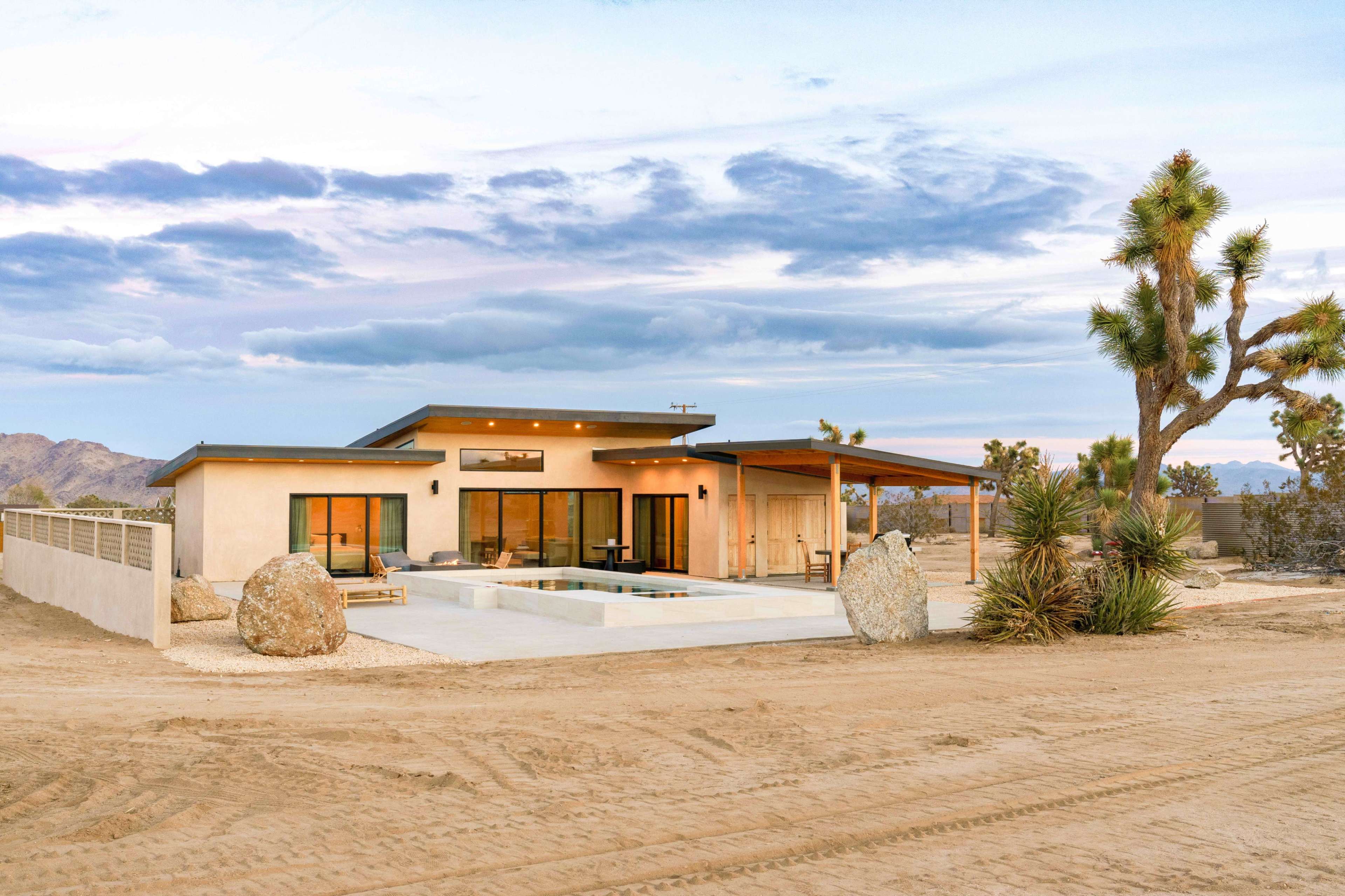 A modern home with large windows and a patio is set against a desert landscape featuring Joshua trees and rocky terrain.