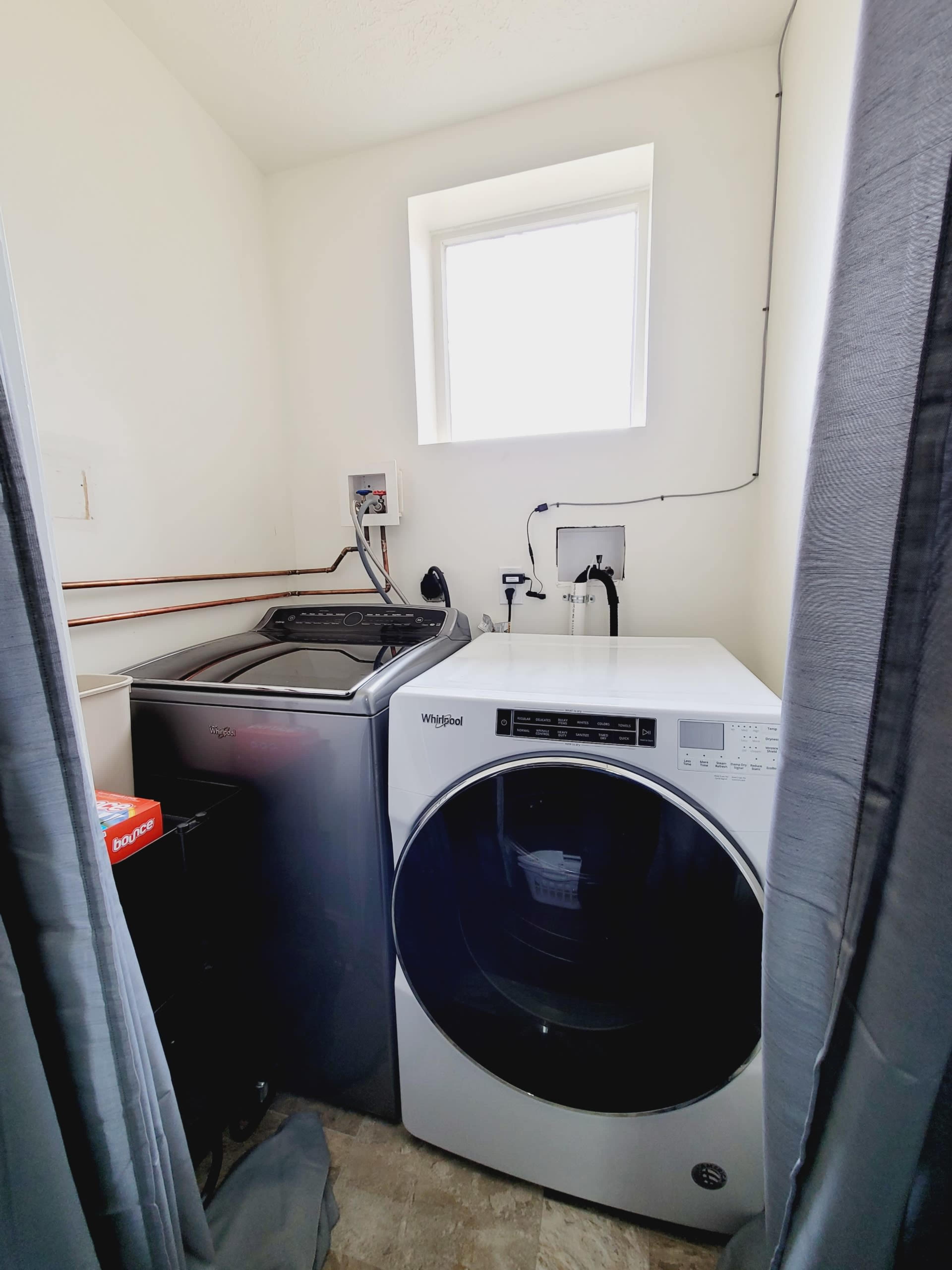 A laundry room with a washing machine and a dryer next to each other, a small window providing natural light, and utility connections on the wall.