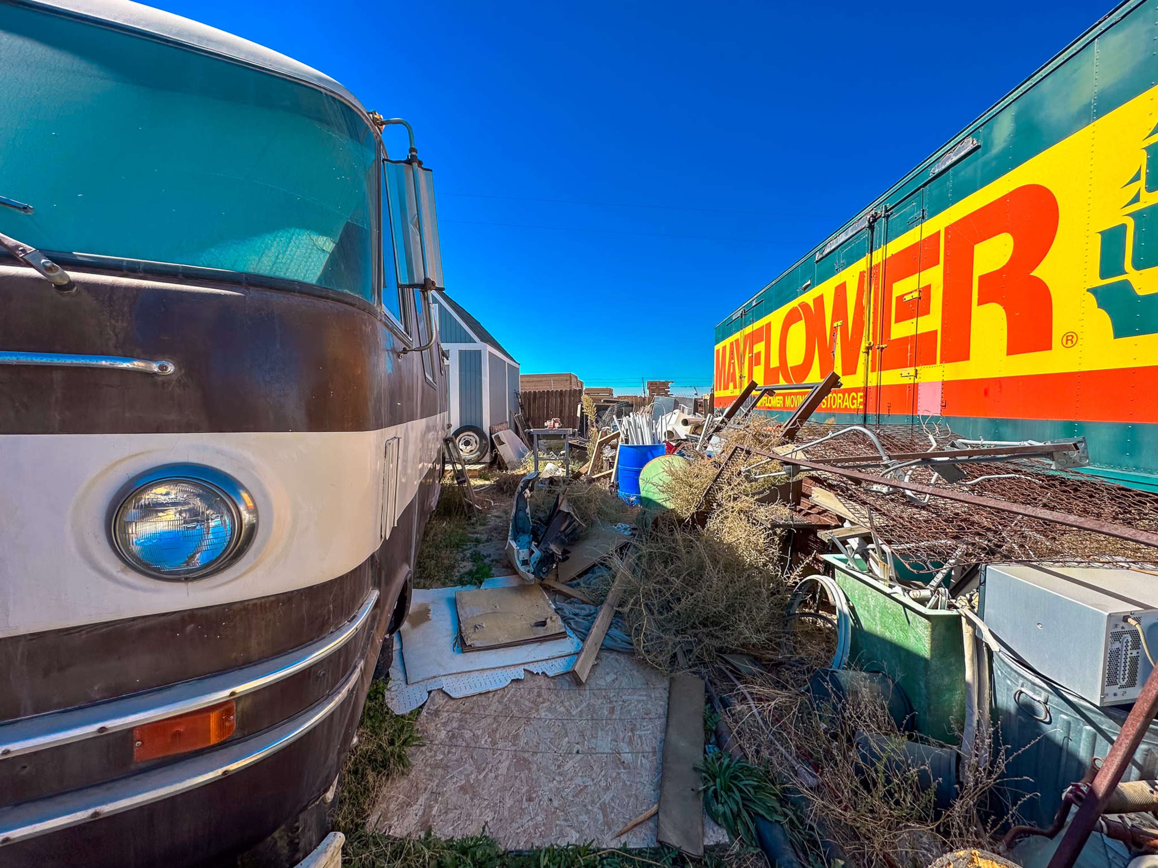 The image shows a narrow space between a vintage bus and a large yellow and green moving truck, surrounded by various items and overgrown vegetation.