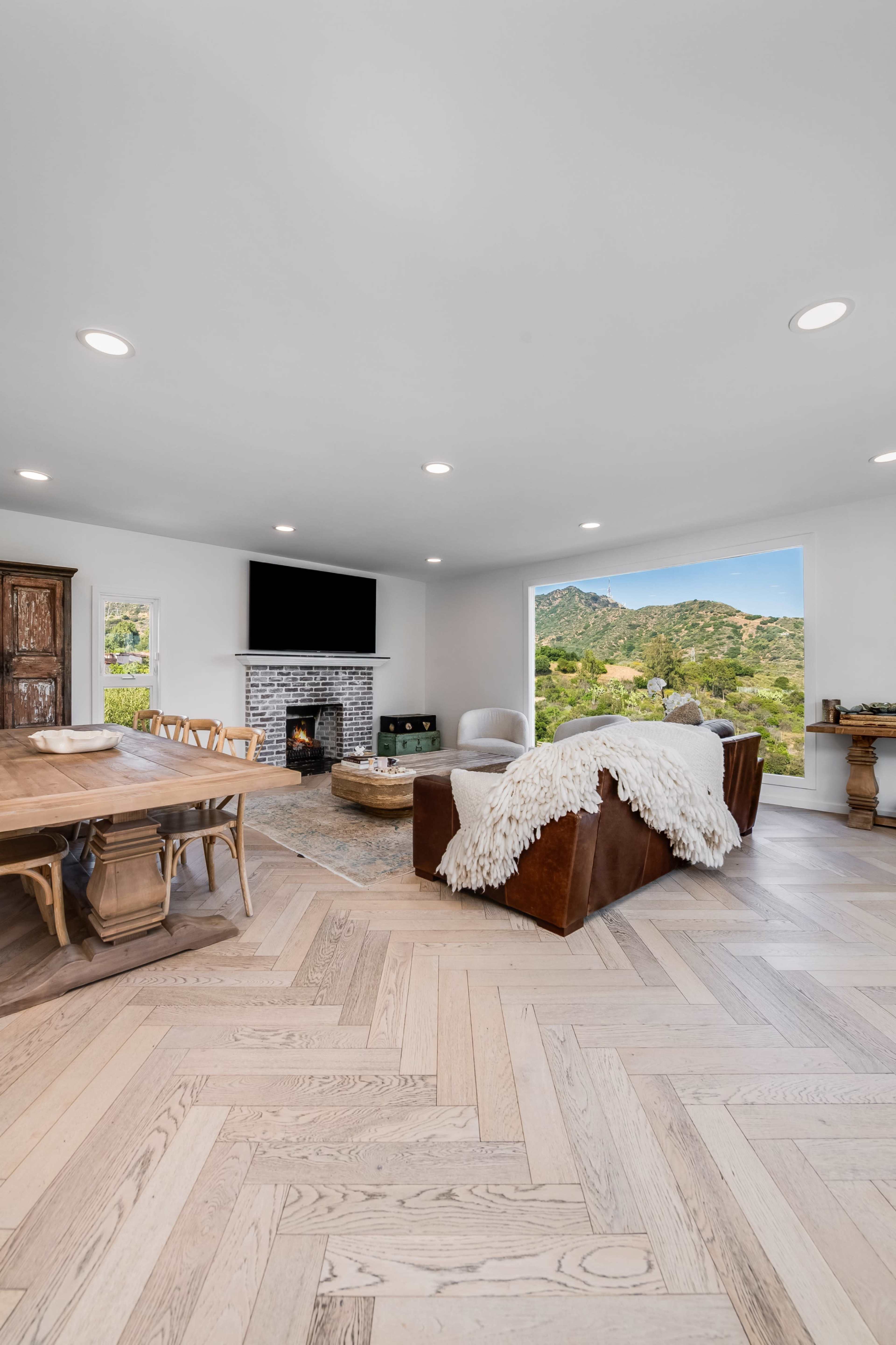 A modern living room features a fireplace, a large window showcasing a scenic view, and a mix of wooden furniture arranged around a light-colored herringbone floor.