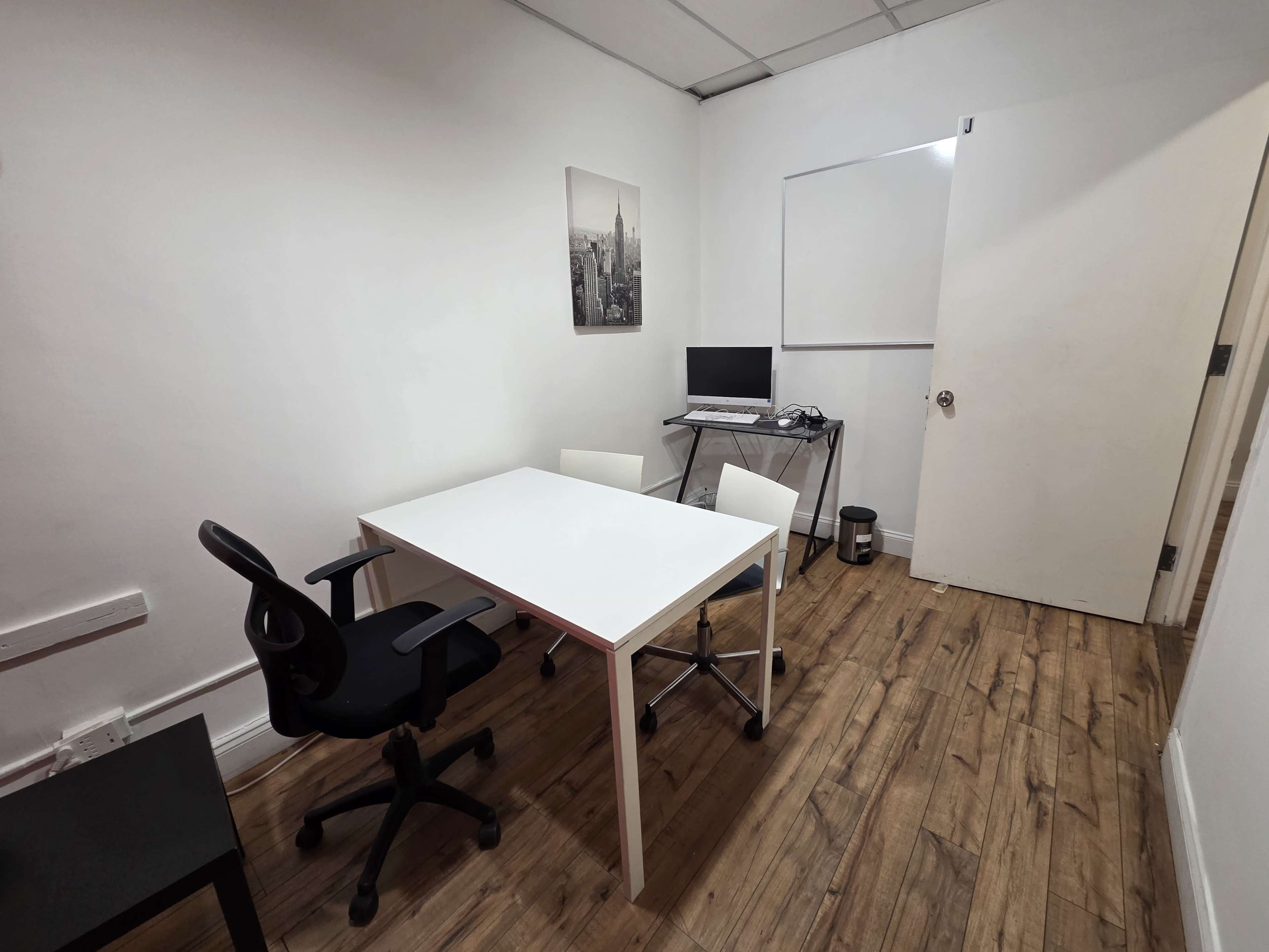 The image shows a small, minimally furnished office with a white table, two chairs, a computer setup, and a large whiteboard on the wall.