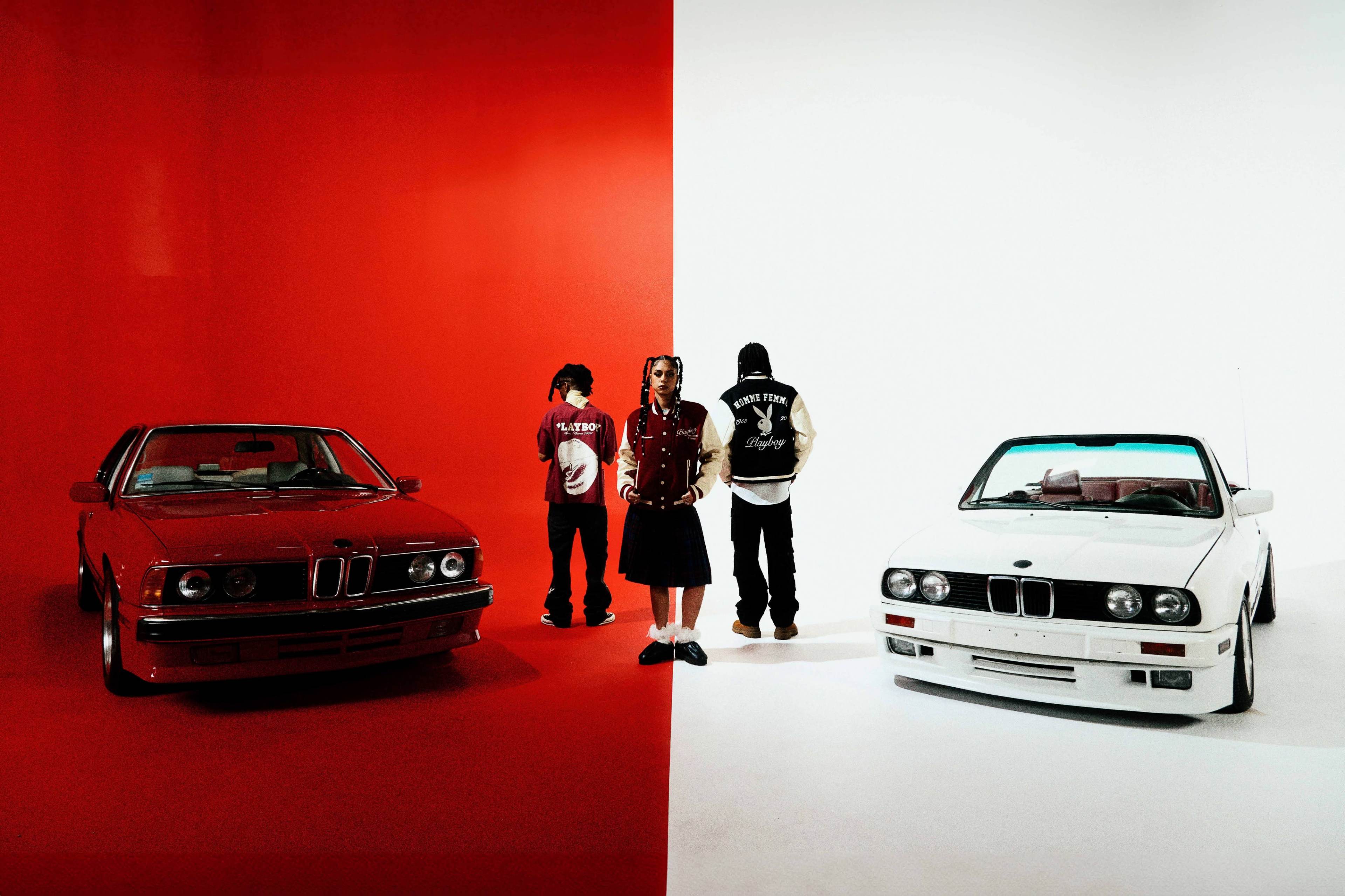 Two BMW cars, one red and one white, are positioned on opposite sides of a stark red and white background, with three individuals standing between them.