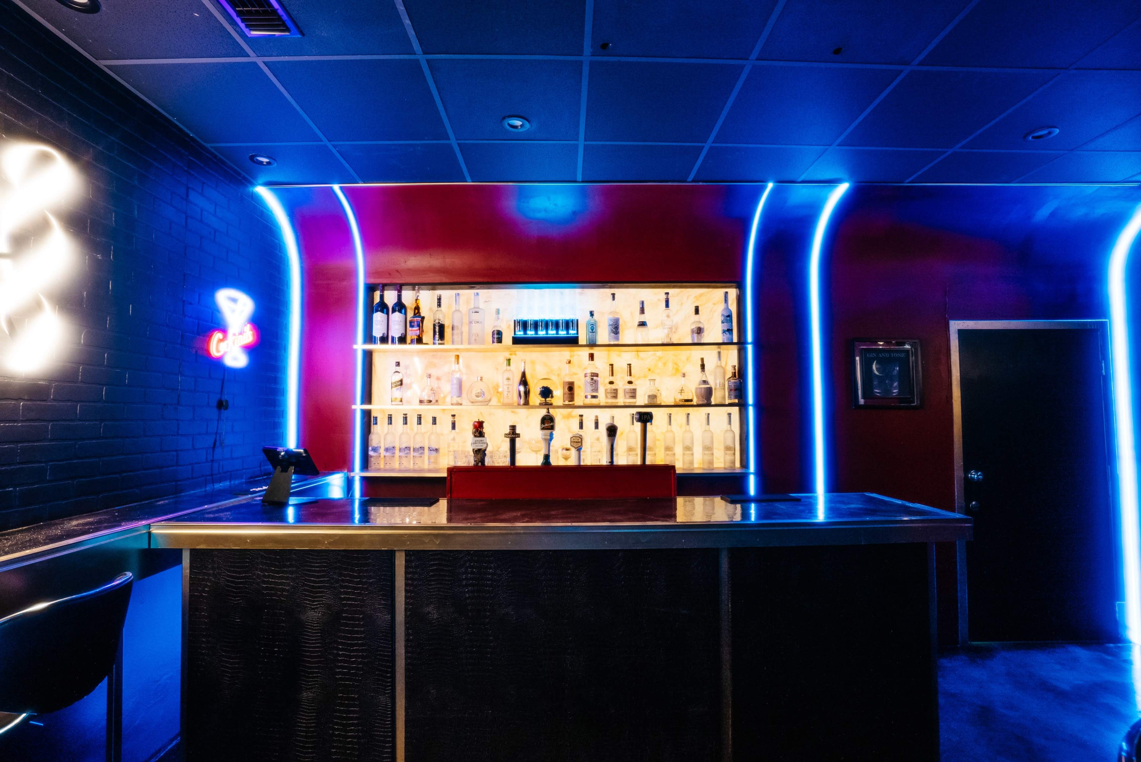 A bar with illuminated shelves displaying various bottles is set against a backdrop of red and black walls.