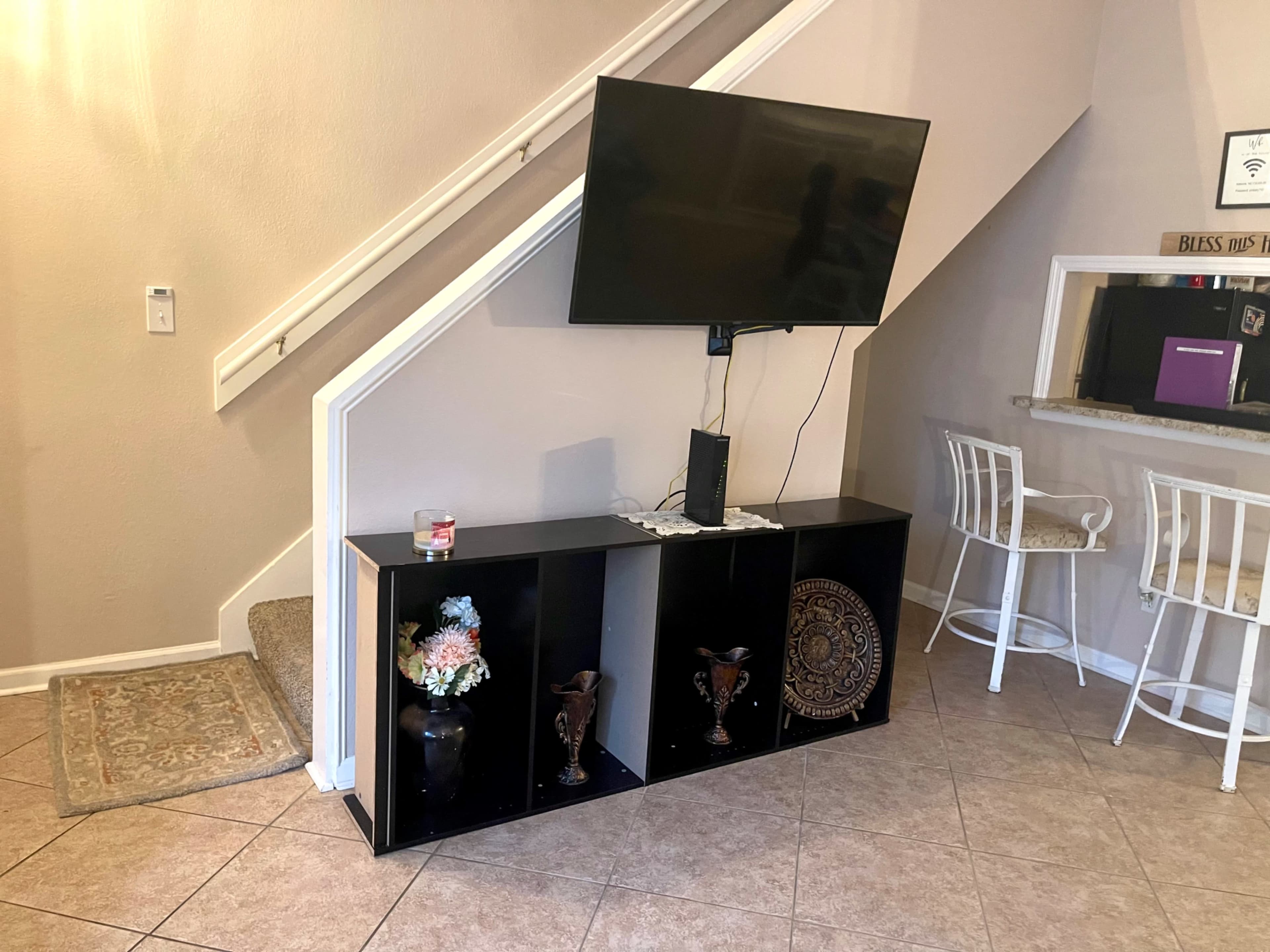 A corner of a room featuring a television mounted on the wall above a black shelving unit with decorative items, positioned next to a staircase.