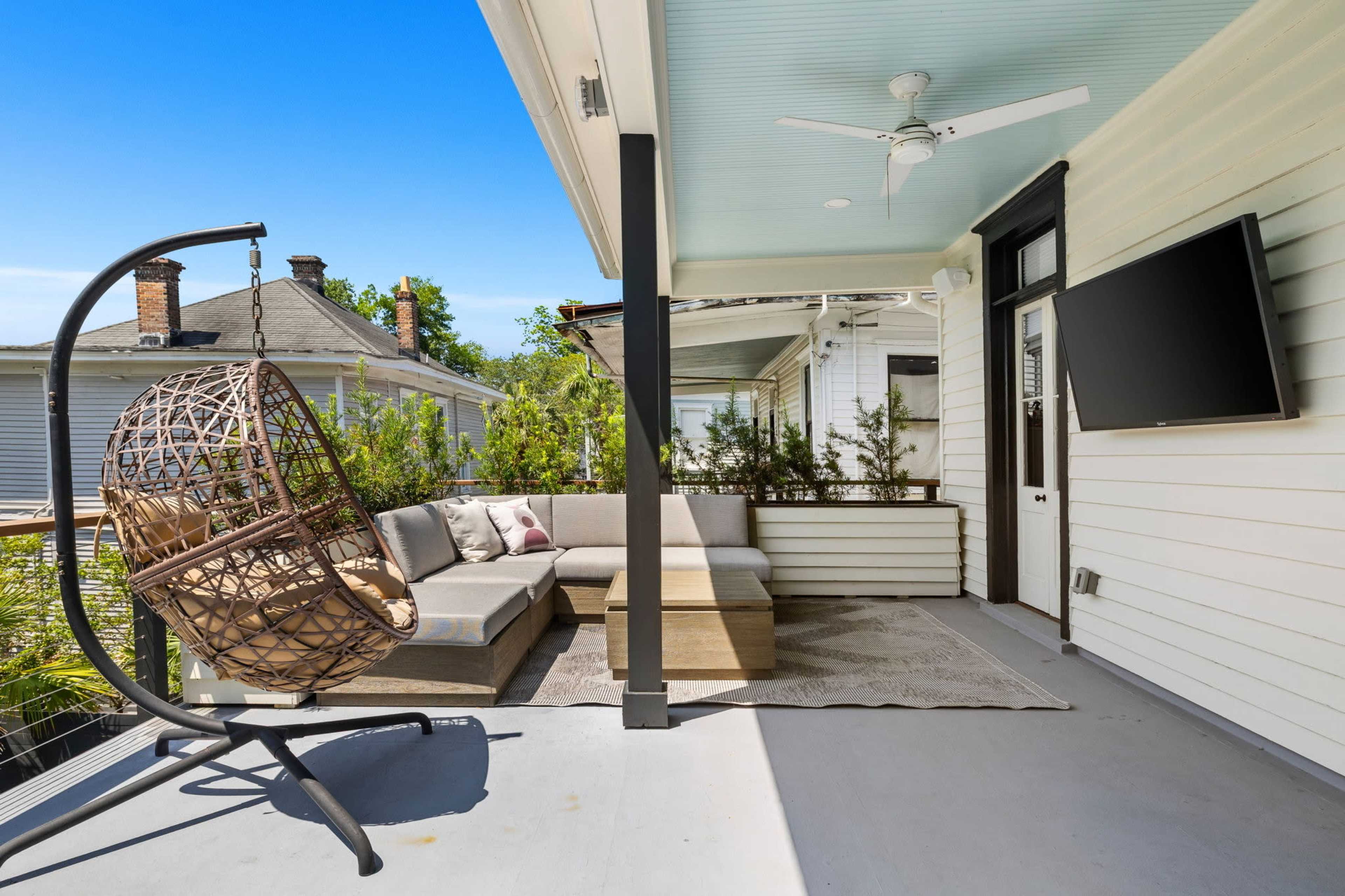 The image features a covered porch area with a hanging chair, a sectional sofa, and a wall-mounted TV.