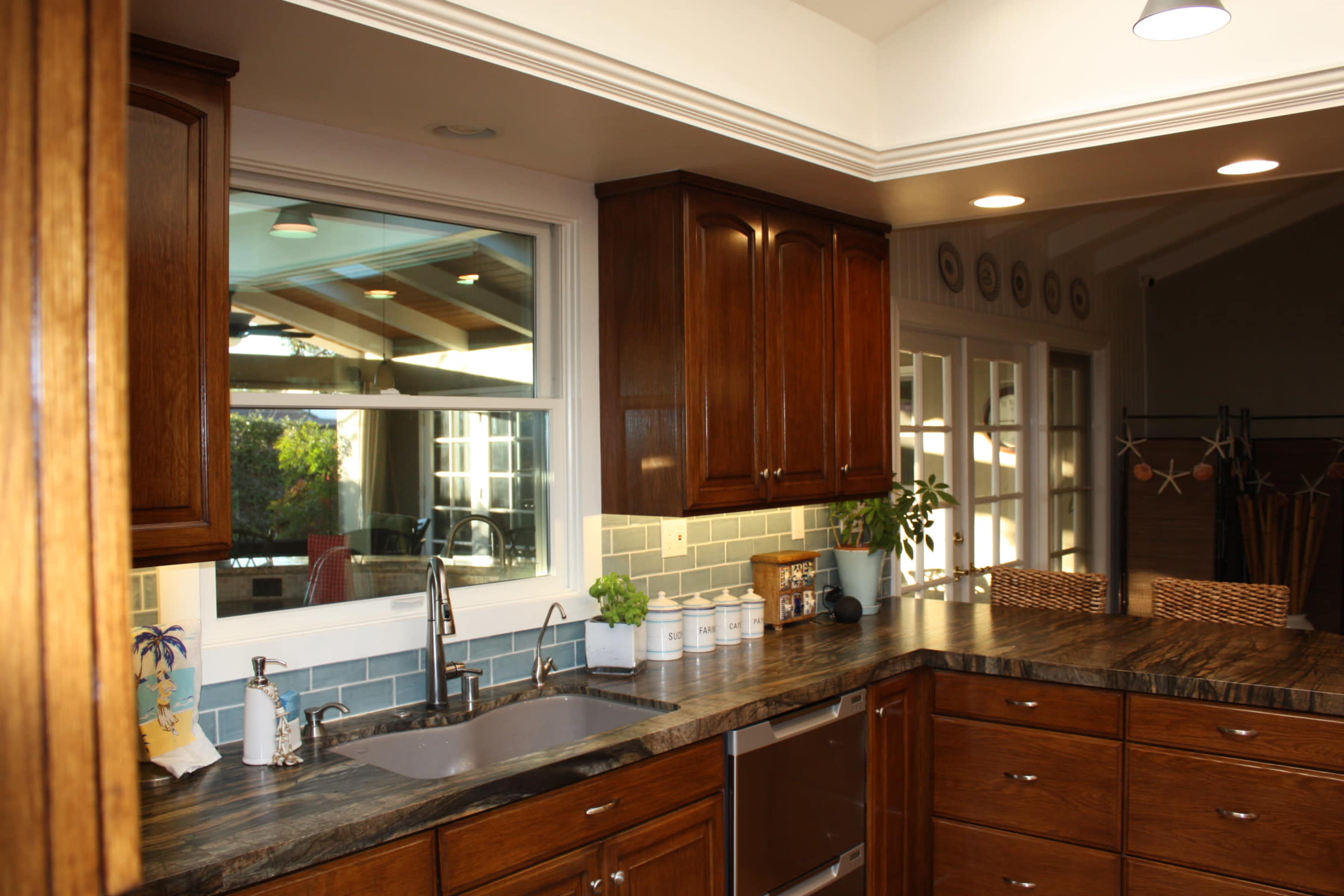 The image shows a modern kitchen with wooden cabinets, a sink under a window, and a granite countertop featuring decorative jars and plants.