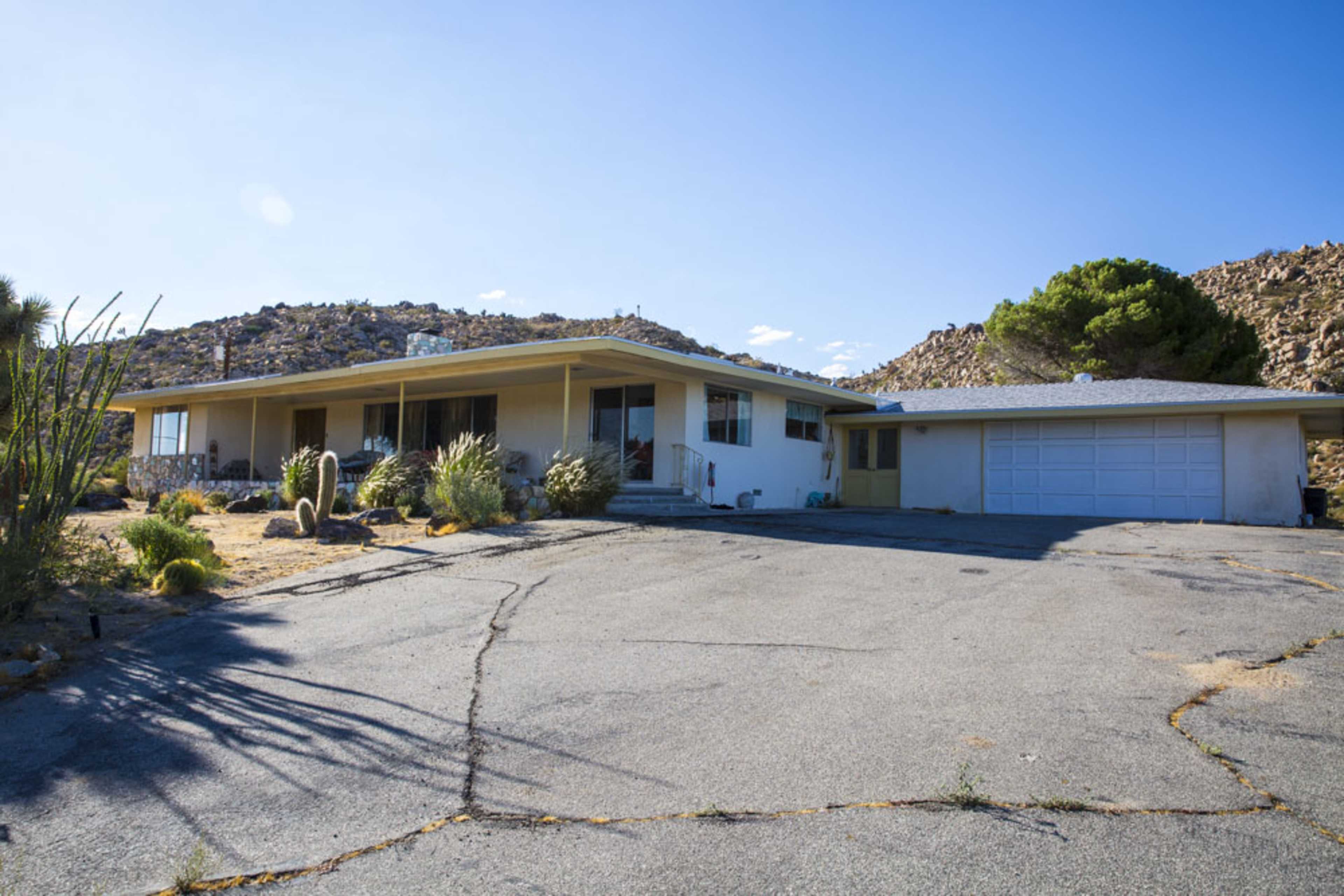 A single-story house with a flat roof is situated on a paved driveway against a backdrop of rocky terrain and desert vegetation.
