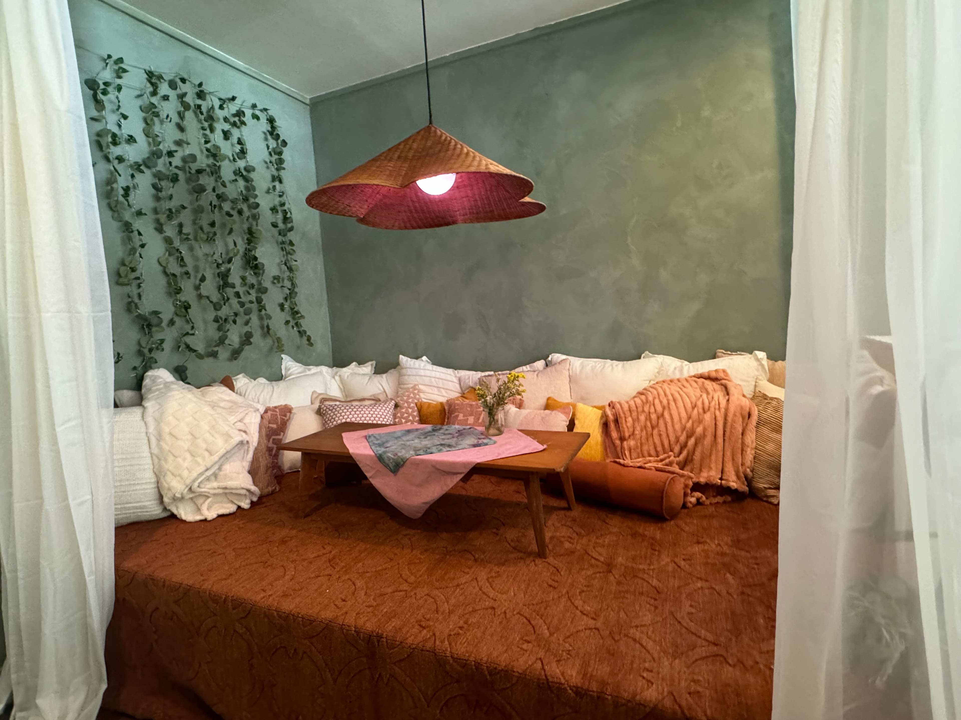 A cozy seating area features a low wooden table surrounded by cushions on a textured orange bedspread, with a hanging light and green wall decor.