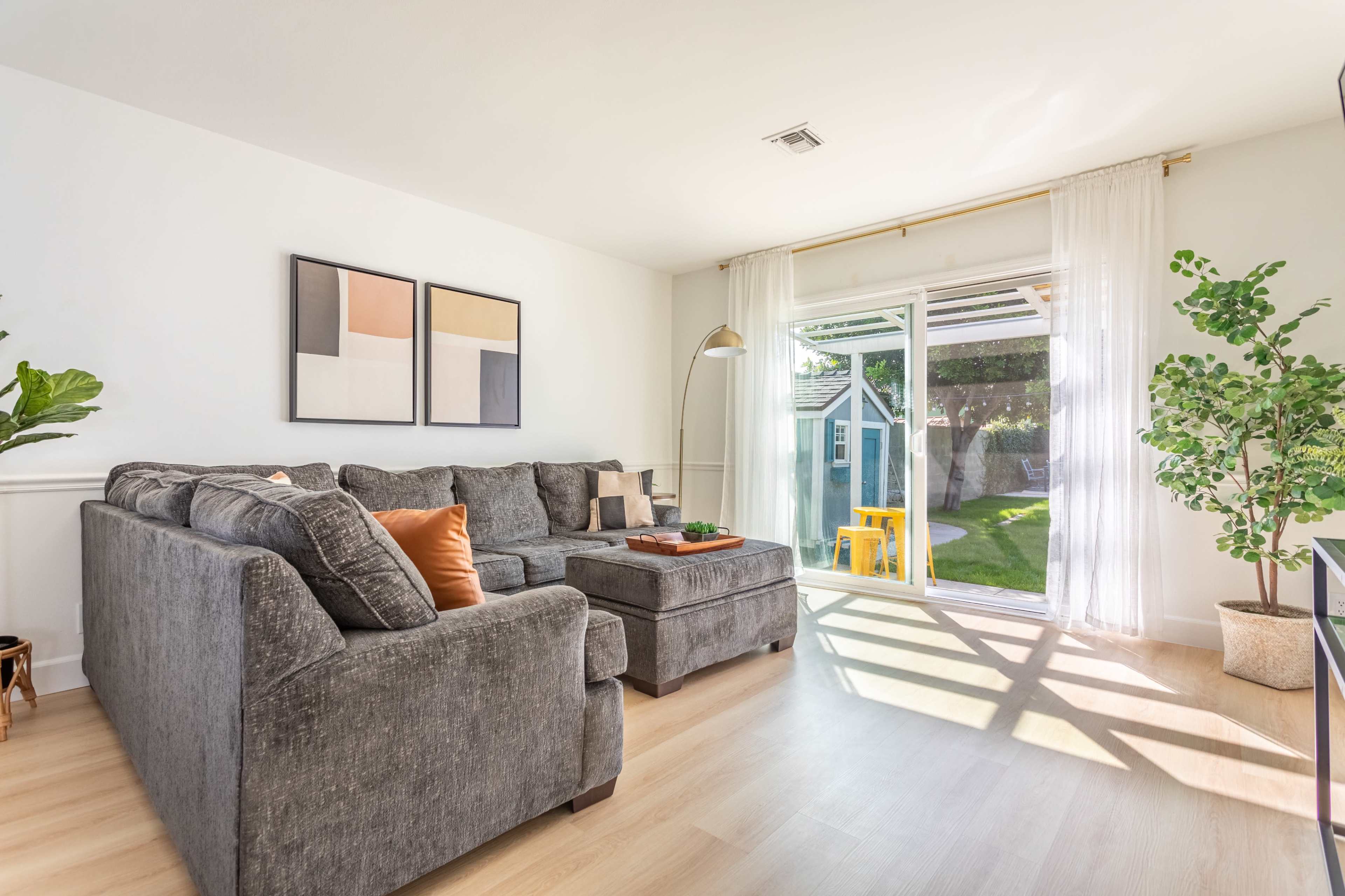 The image shows a modern living room featuring a grey sectional sofa, large windows with sheer curtains, and a view of a backyard.