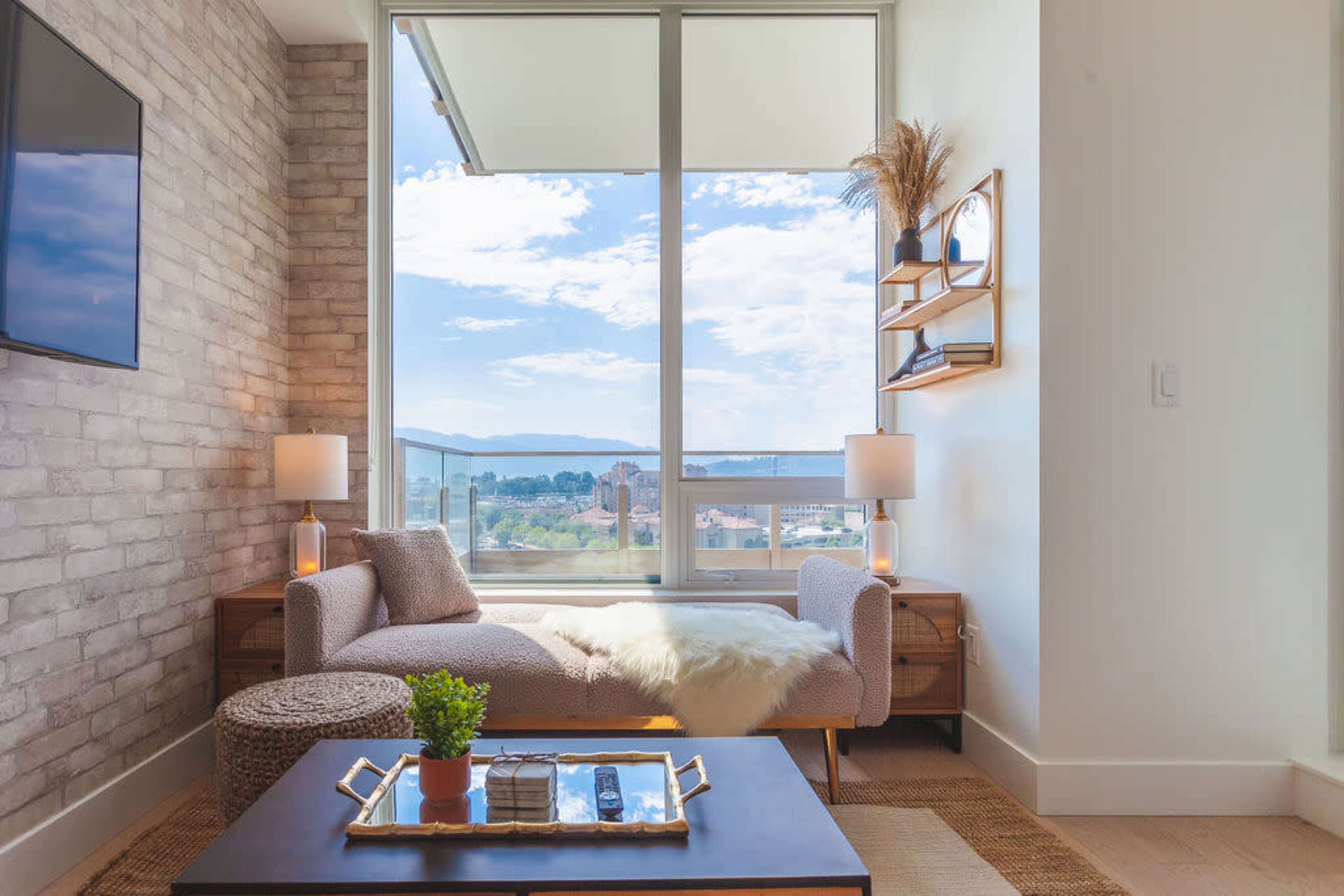 A well-lit living area features a sofa by a large window with a view, complemented by side tables, lamps, and decorative elements.