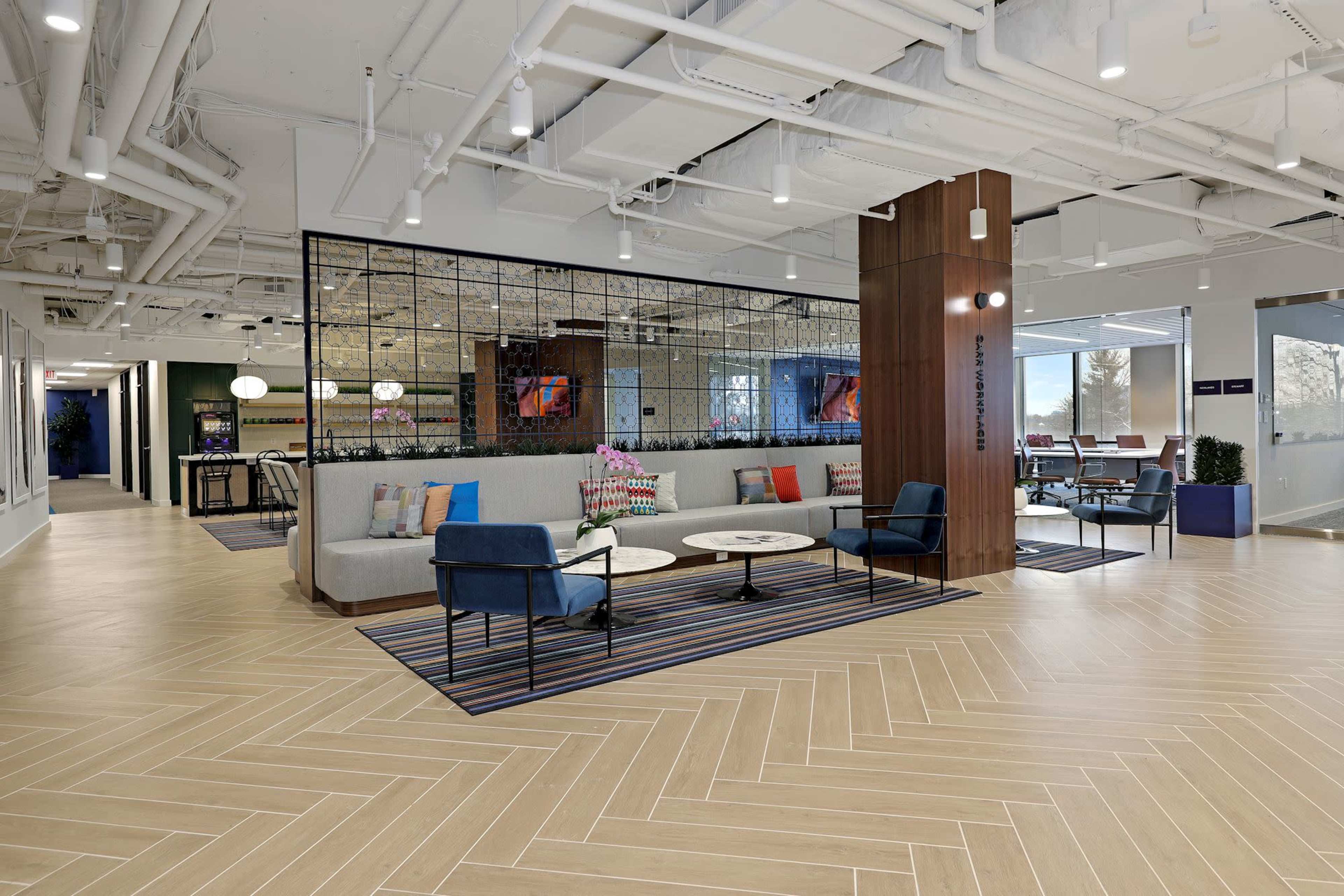 The image shows a modern office lounge area with comfortable seating, a glass partitioned meeting space, and wooden floors.
