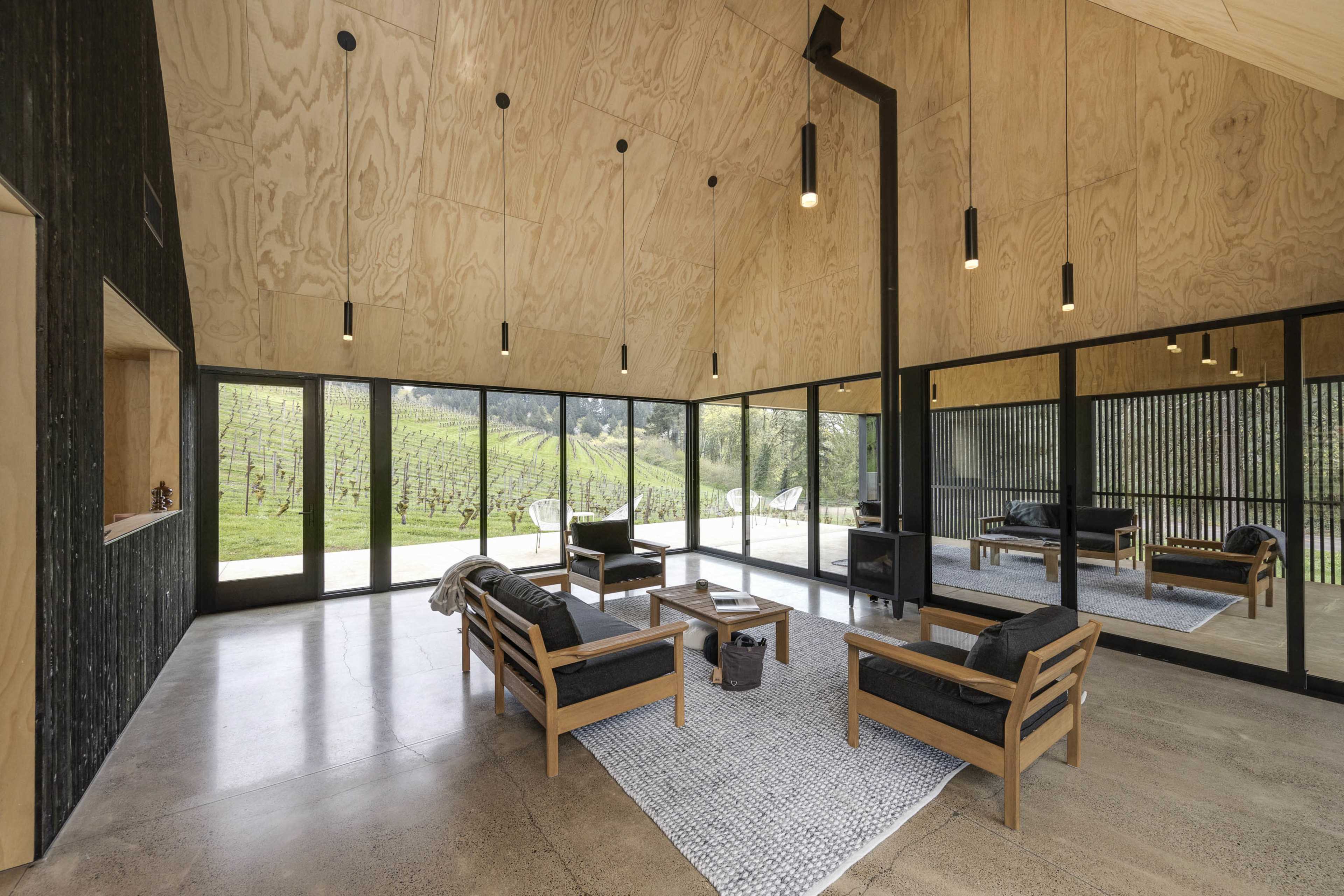 The image shows a modern, open-concept living area with large windows overlooking a vineyard, featuring wooden furniture and a central fireplace.