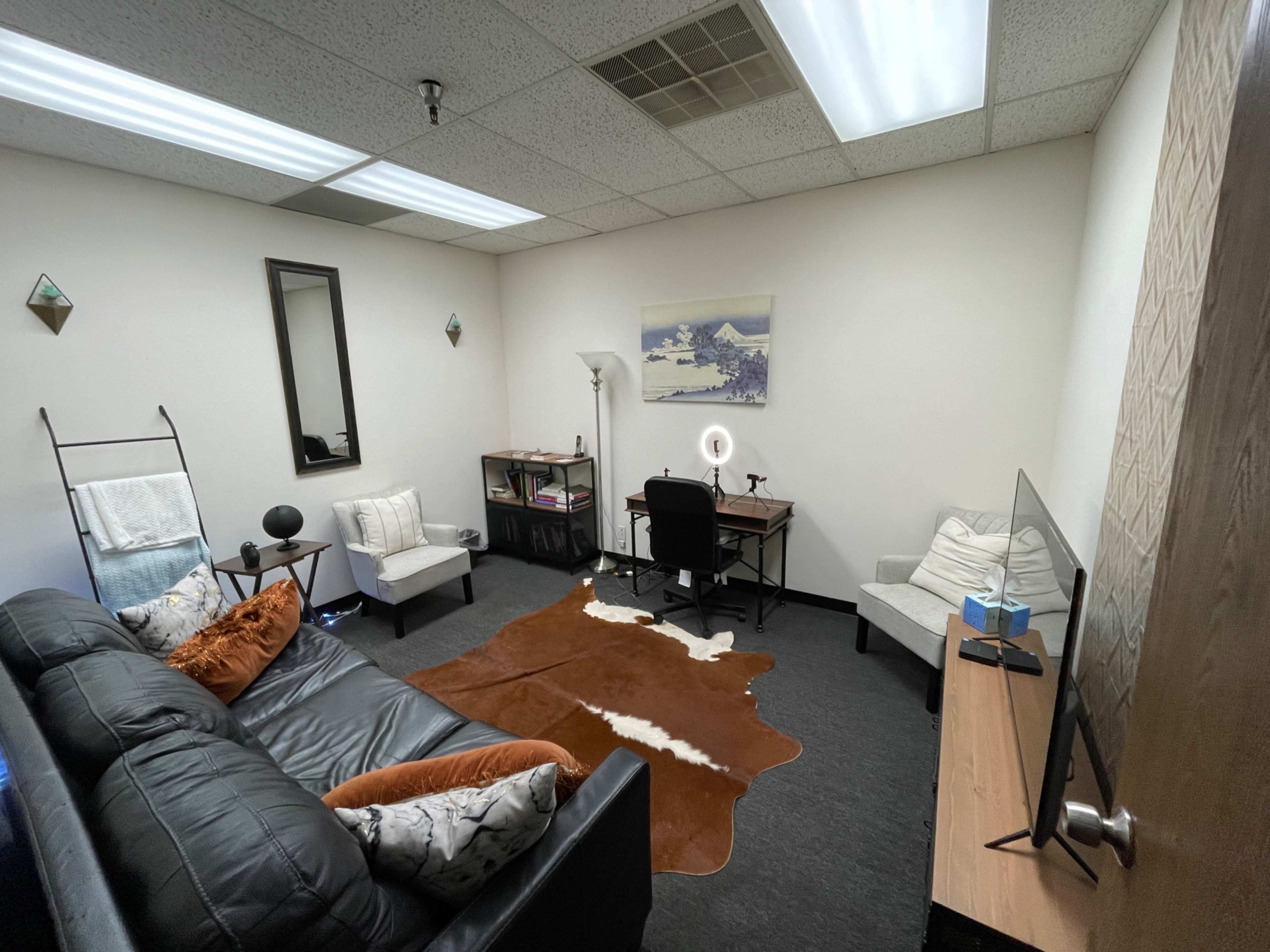 A small office space featuring a black couch, a desk with a chair, and decorative elements like a mirror and a painting on the wall.