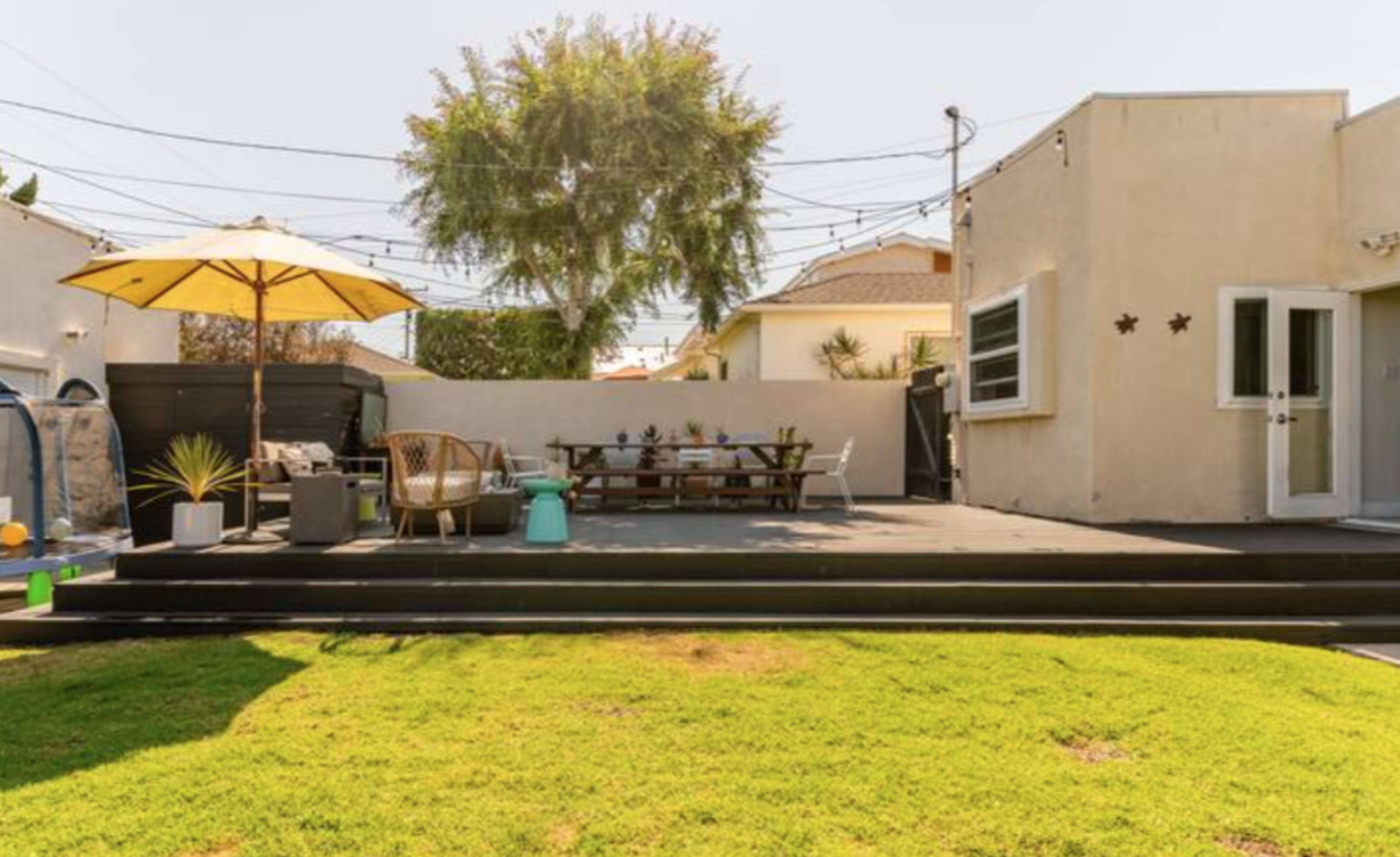 The image shows a backyard featuring a patio with seating, a dining table under an umbrella, and a well-maintained lawn.