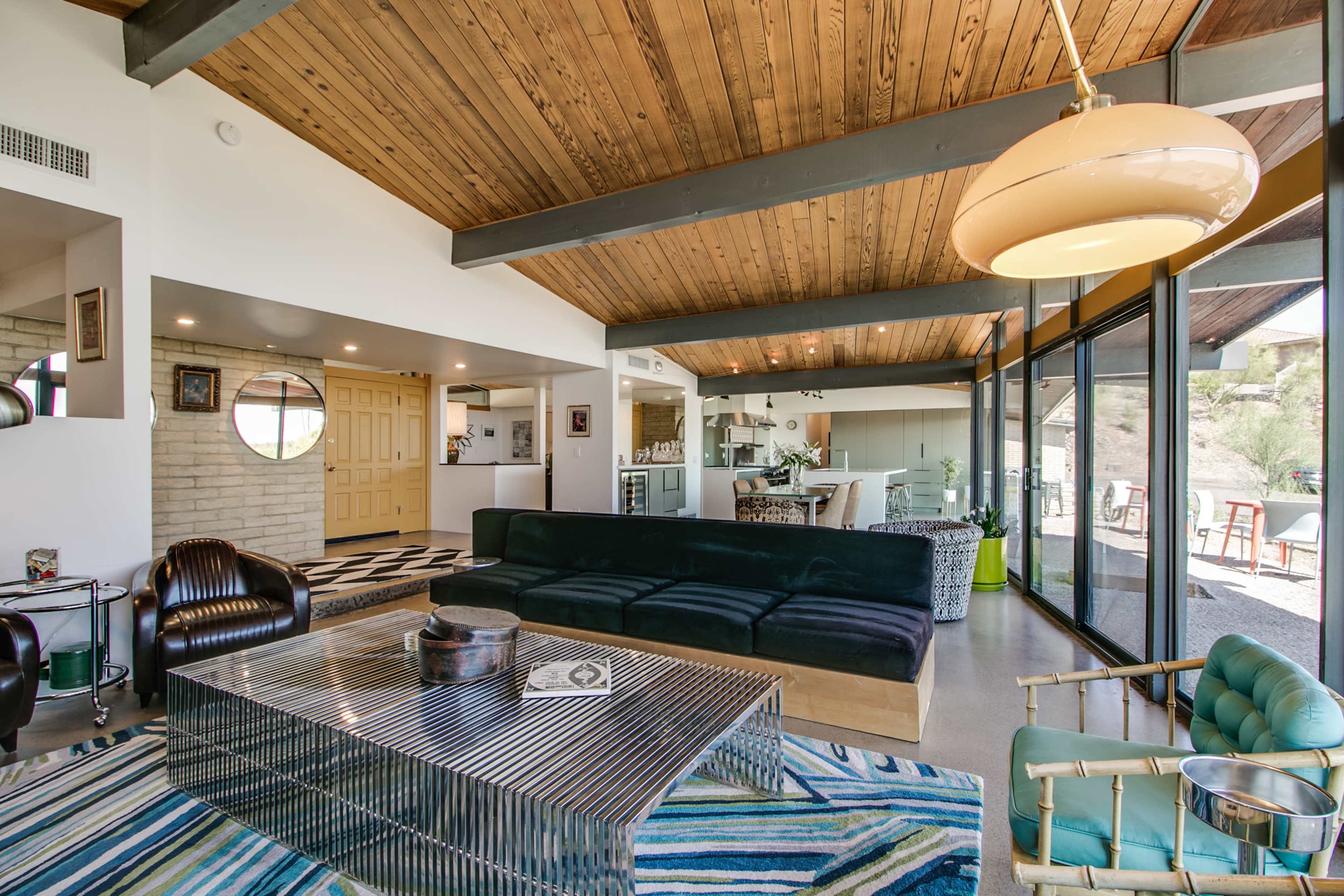 A modern living room with a wooden ceiling, a sectional sofa, a glass coffee table, and large windows overlooking an outdoor space.