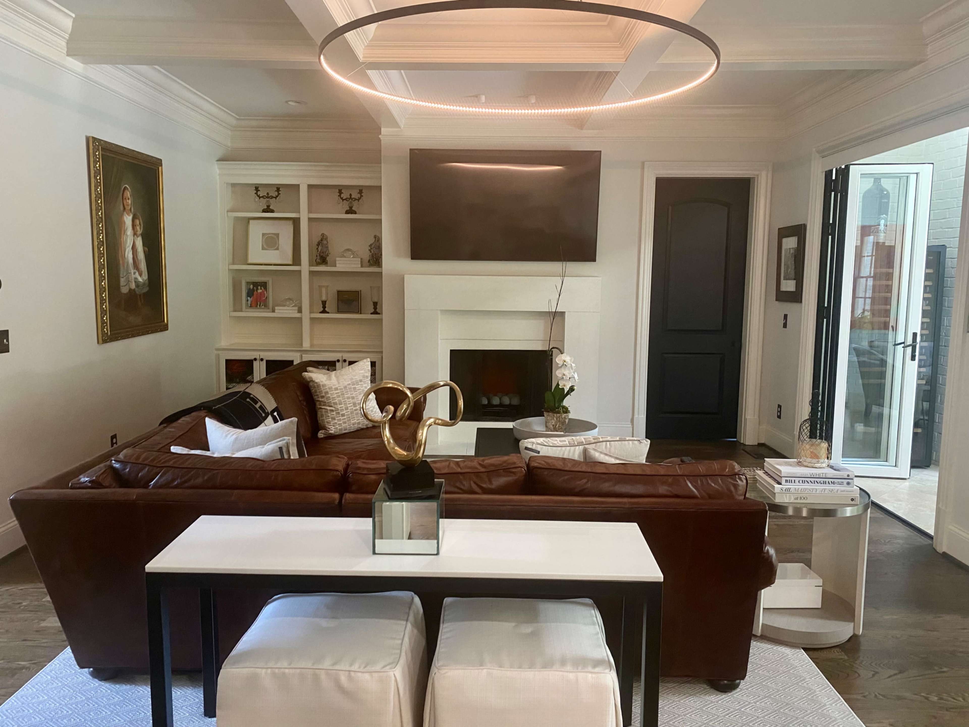 A modern living room with a brown leather sectional sofa, a circular ceiling light, a mounted flat-screen TV above a fireplace, and large black-framed doors leading to an outdoor area.