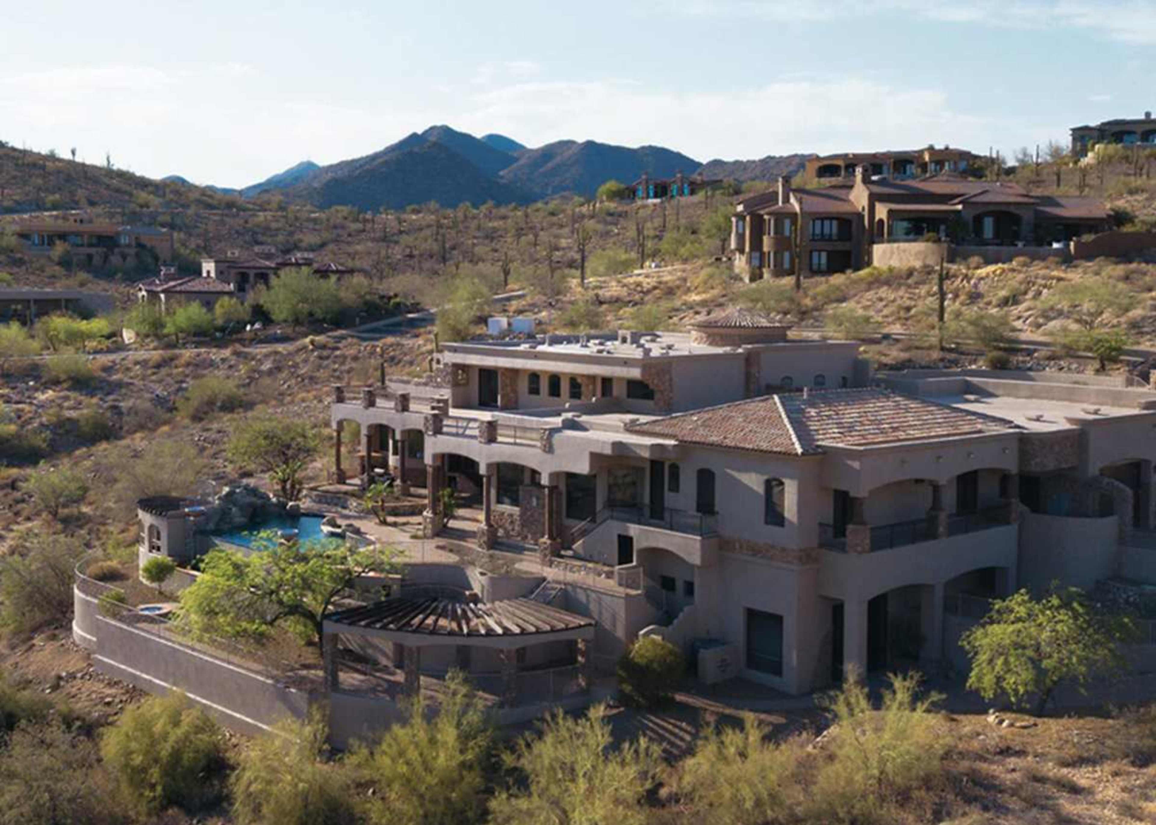 The image shows a large, multi-story house with a swimming pool, situated in a desert landscape surrounded by mountains and sparse vegetation.