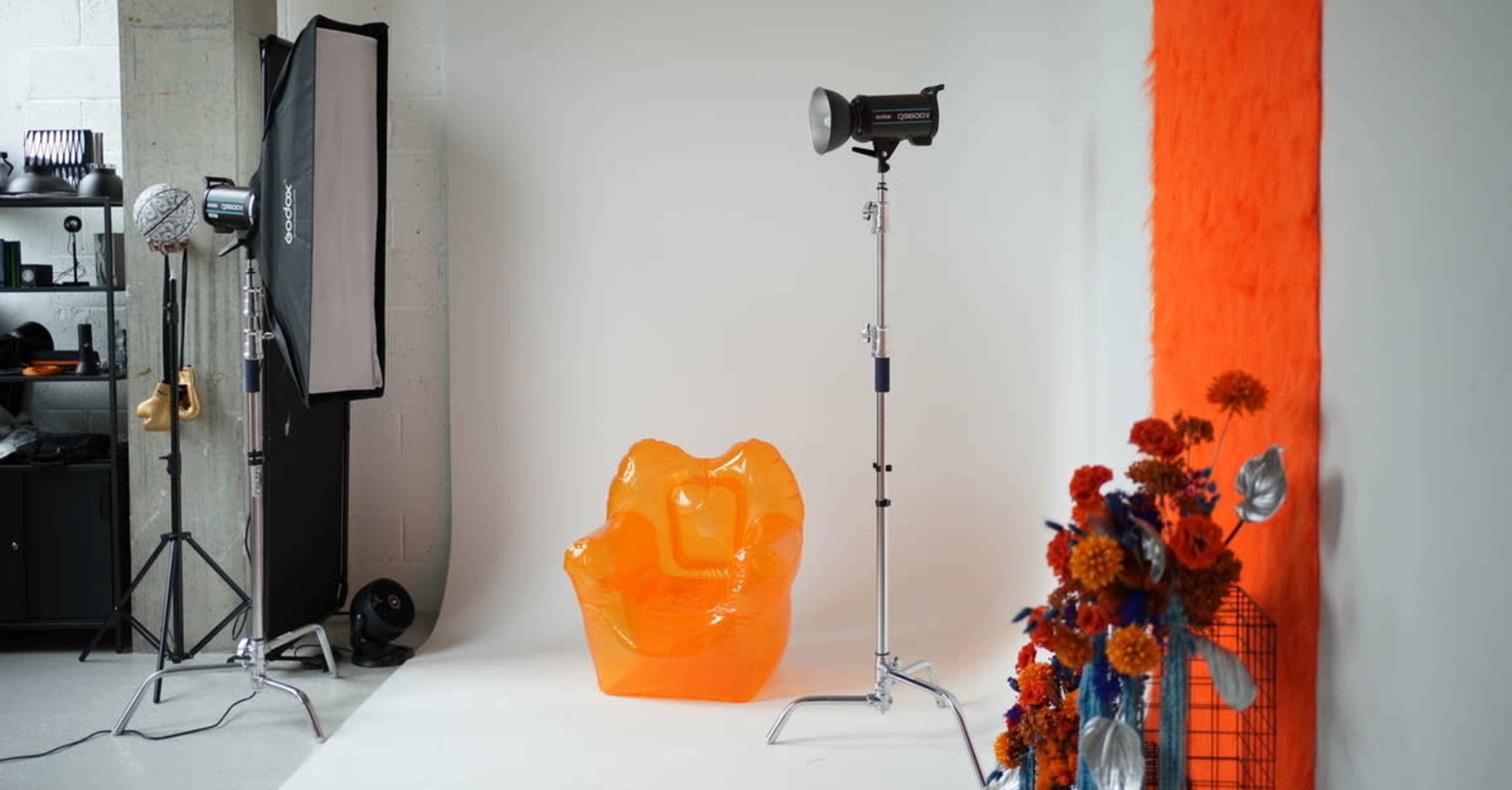 The image shows a photography studio setup featuring an orange transparent chair, a lighting equipment stand, and a bouquet of flowers against a light background.