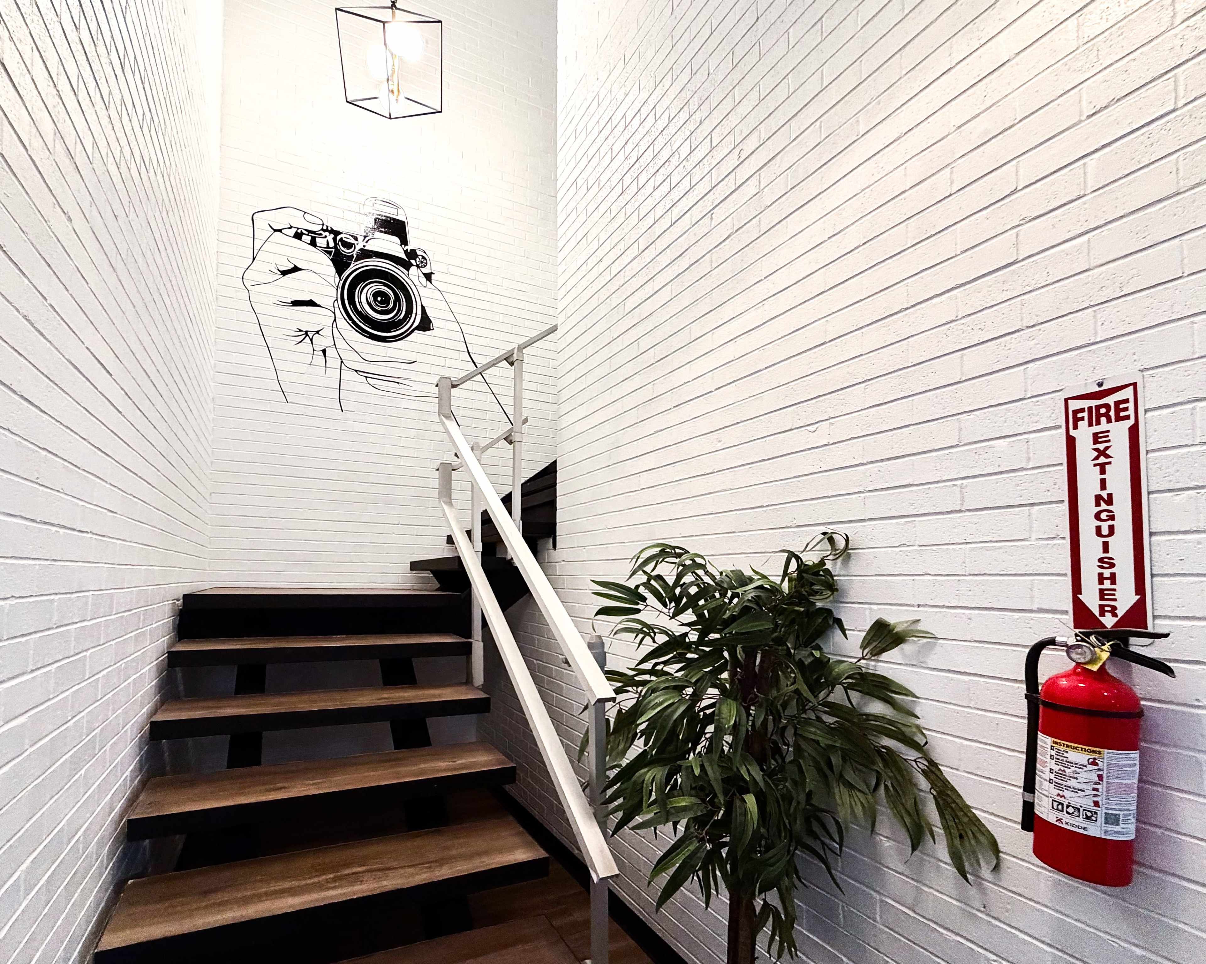 A staircase leads upward alongside a white-brick wall featuring a large camera mural, with a fire extinguisher mounted nearby and a potted plant at the base.