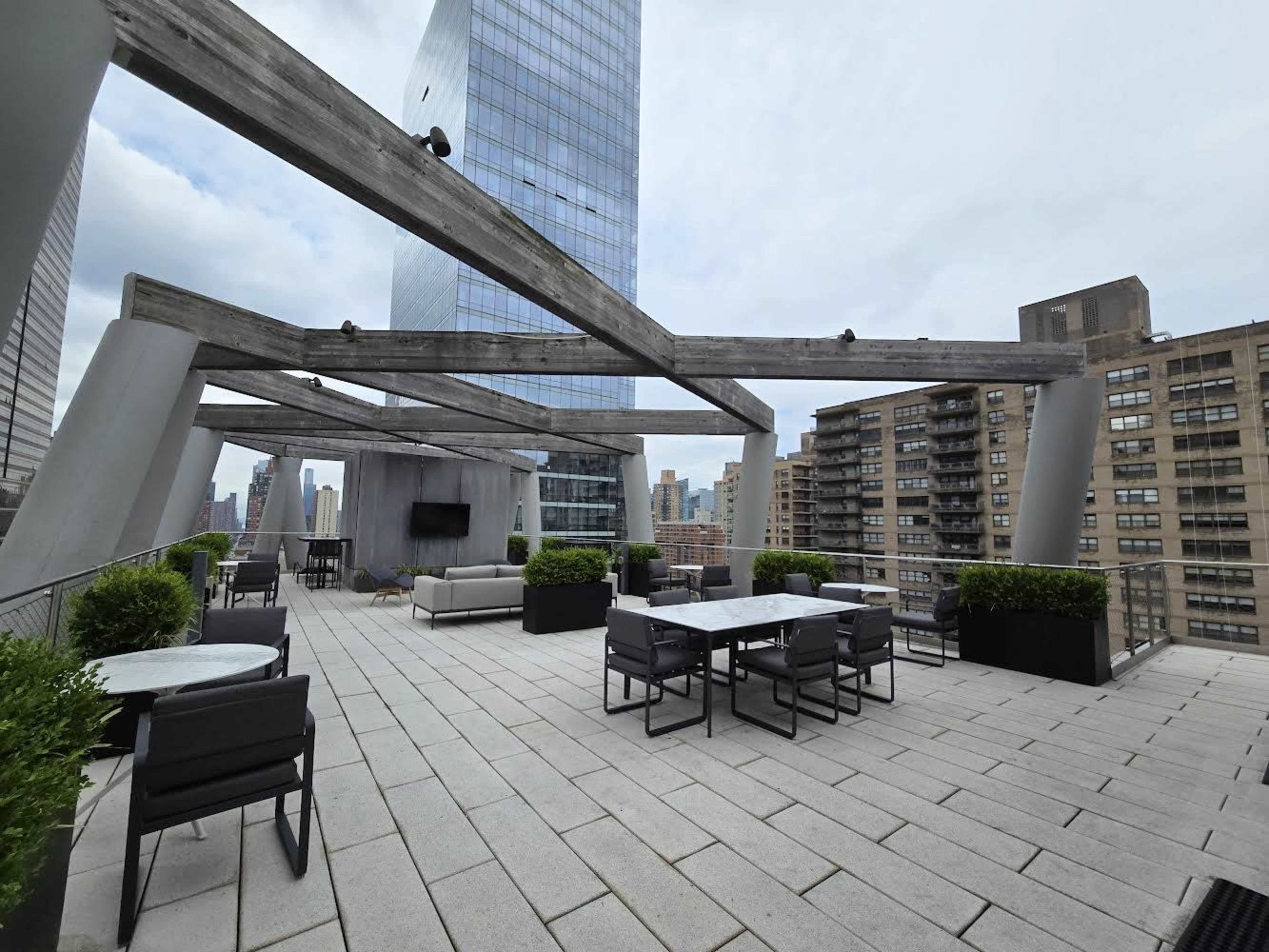 A spacious rooftop terrace featuring modern furniture, planters, and a backdrop of tall buildings and a glass skyscraper against a cloudy sky.
