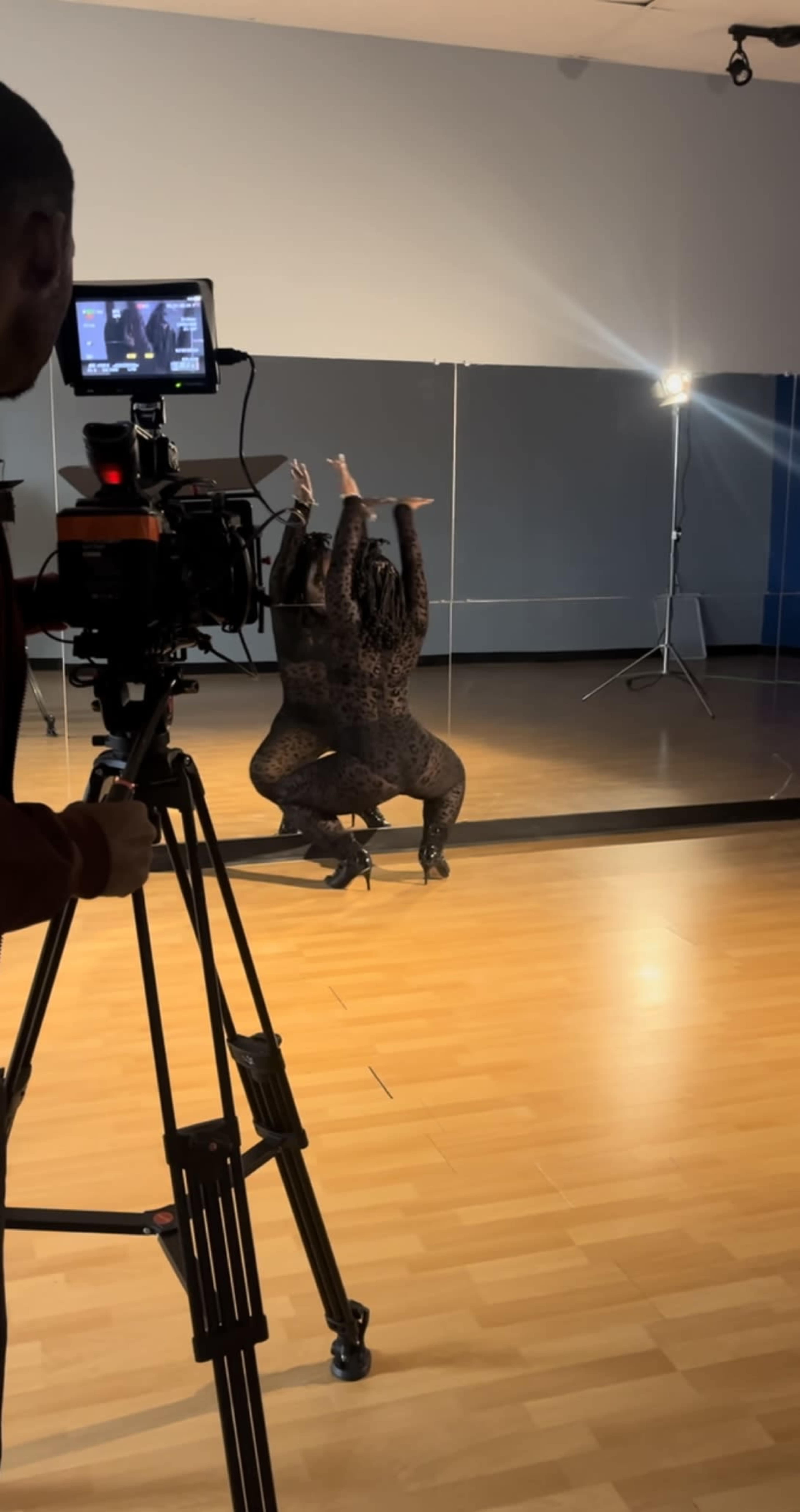 A dancer in a black outfit performs near a mirror while a camera is positioned nearby on a tripod.