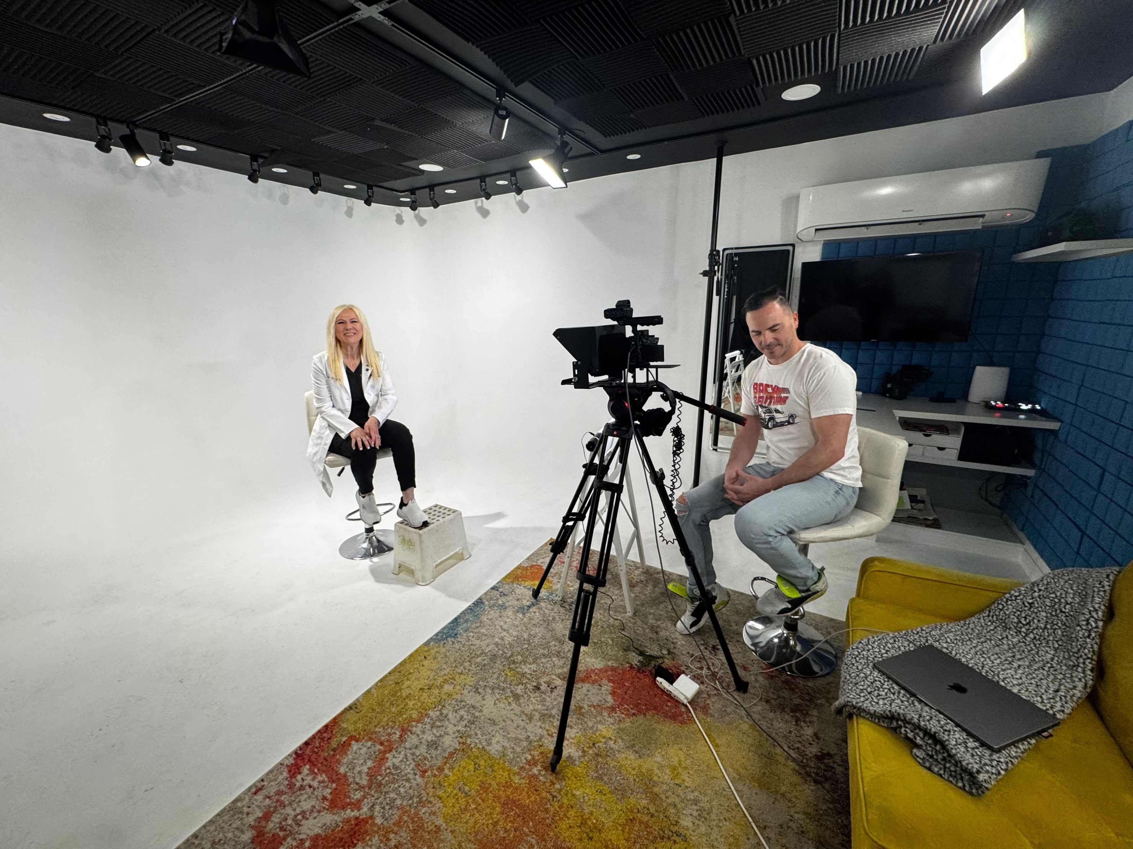 A blonde woman sits on a stool in front of a camera setup in a white studio, while a man adjusts equipment nearby.