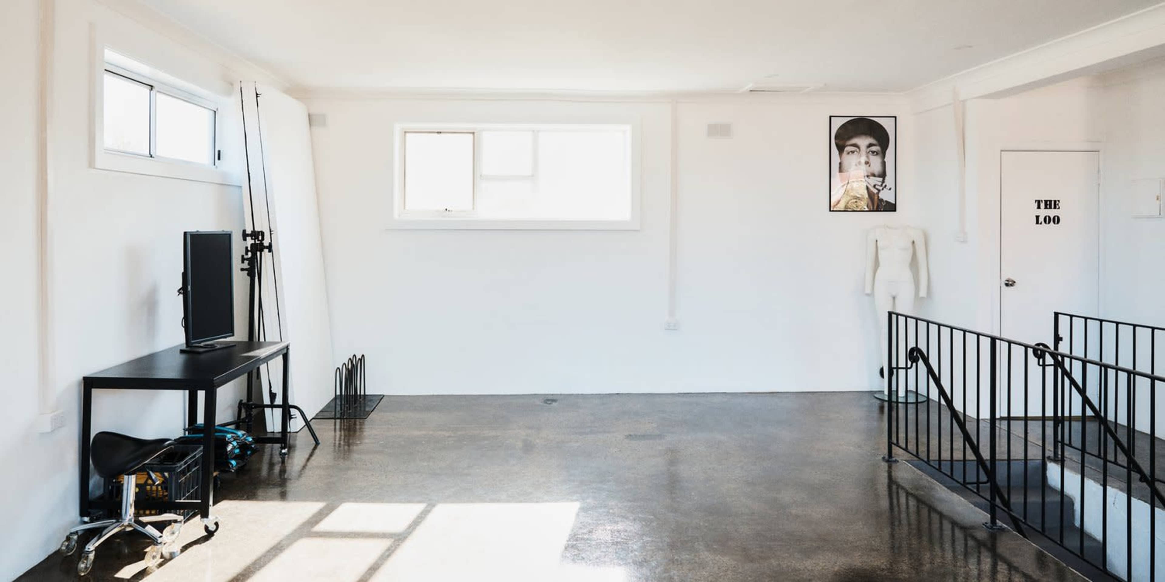 A spacious, empty room with a black table, a window, and a staircase leading to a lower level, featuring a mannequin and a framed photo on the wall.