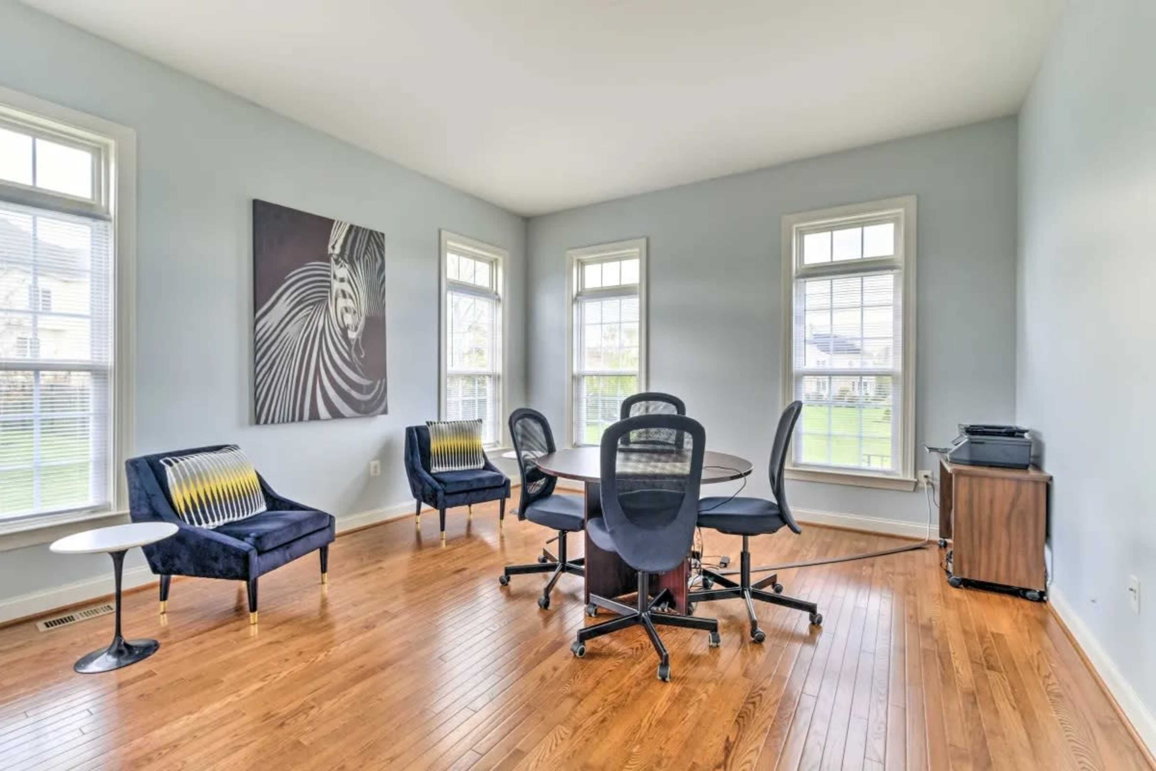A bright, airy meeting room features a round table surrounded by six office chairs, with two blue armchairs and large windows allowing natural light to enter.