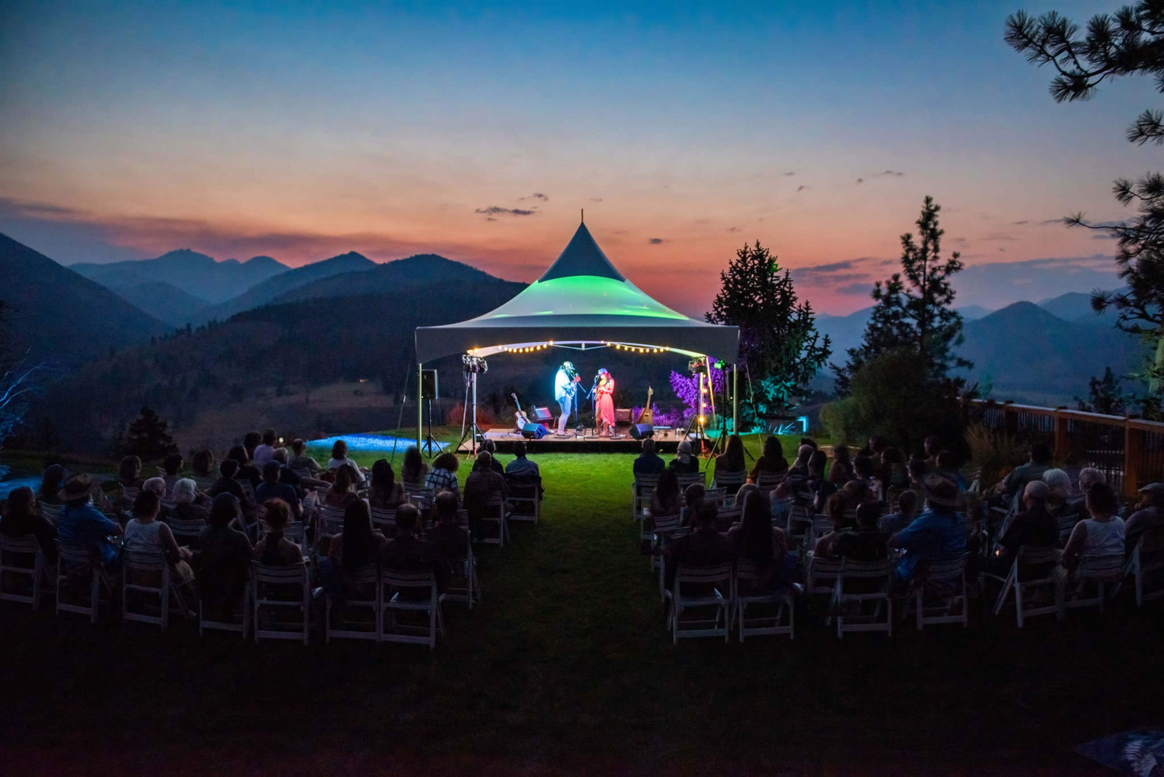 A crowd sits on folding chairs under a tent as performers entertain at dusk, with mountains in the background.