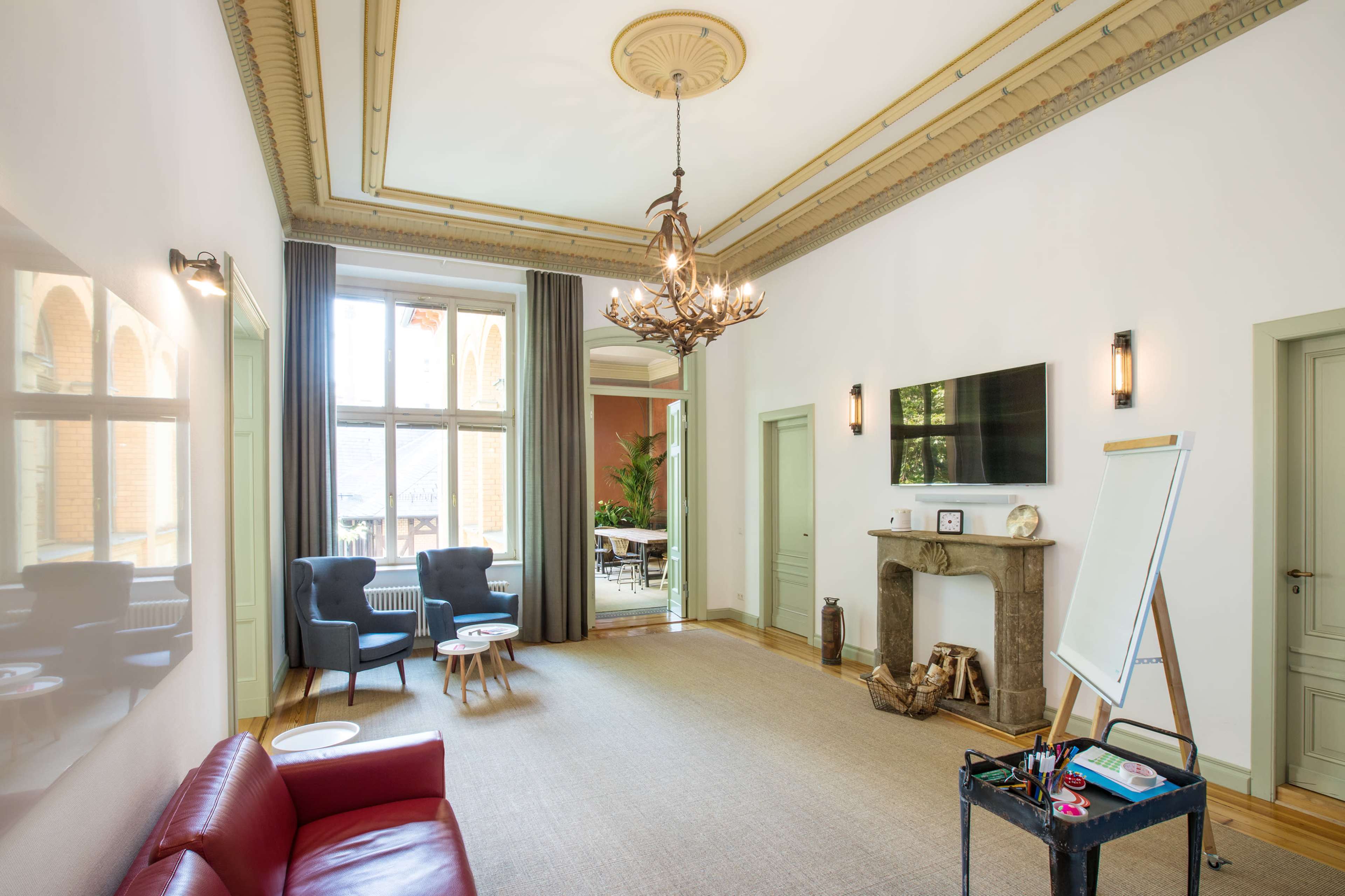 The image shows a spacious, well-lit room featuring a red sofa, two armchairs, a flat-screen TV, a fireplace, and a whiteboard.
