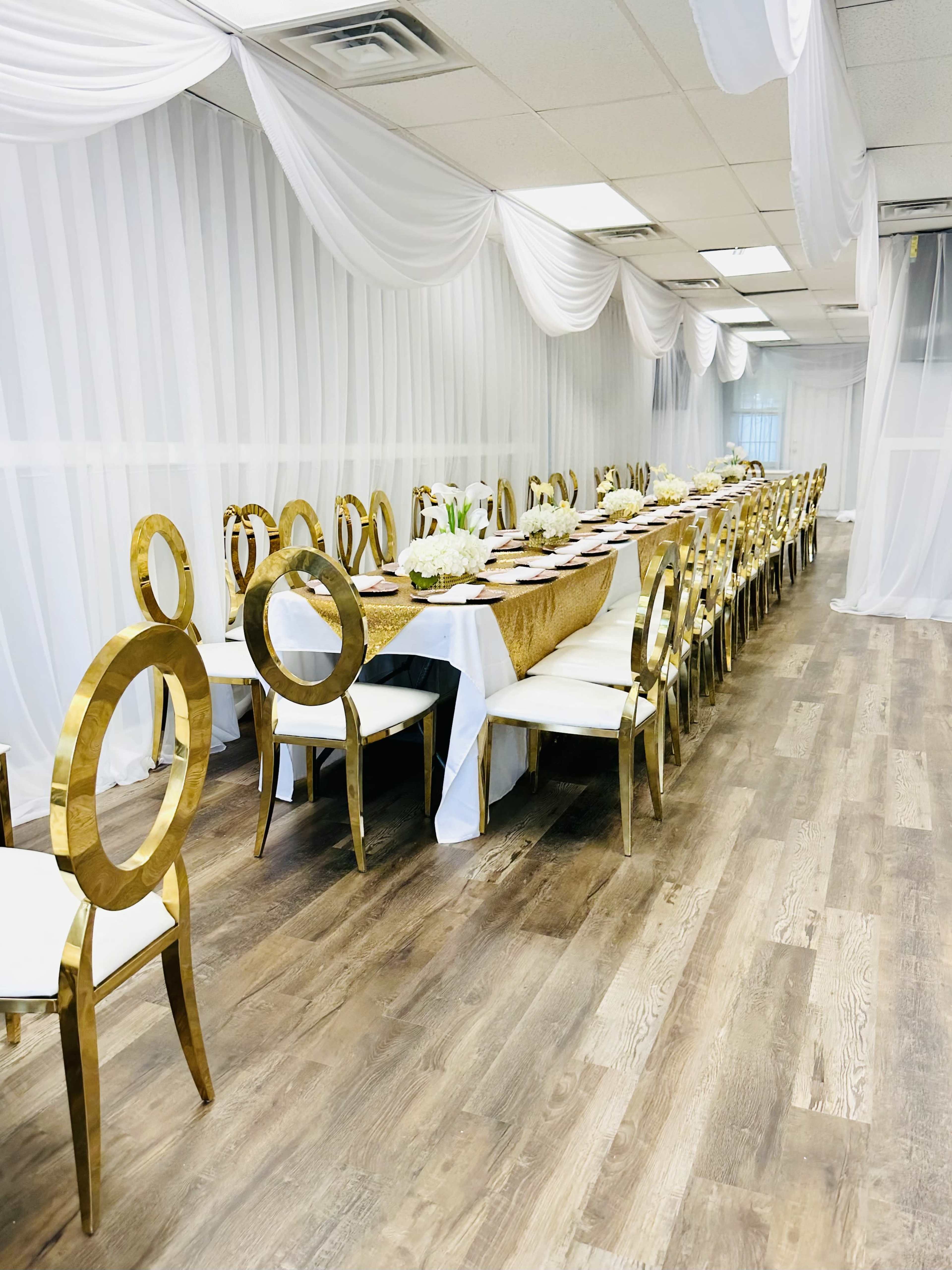 A long table is set with elegant decor and surrounded by gold chairs in a brightly lit room with white drapery.
