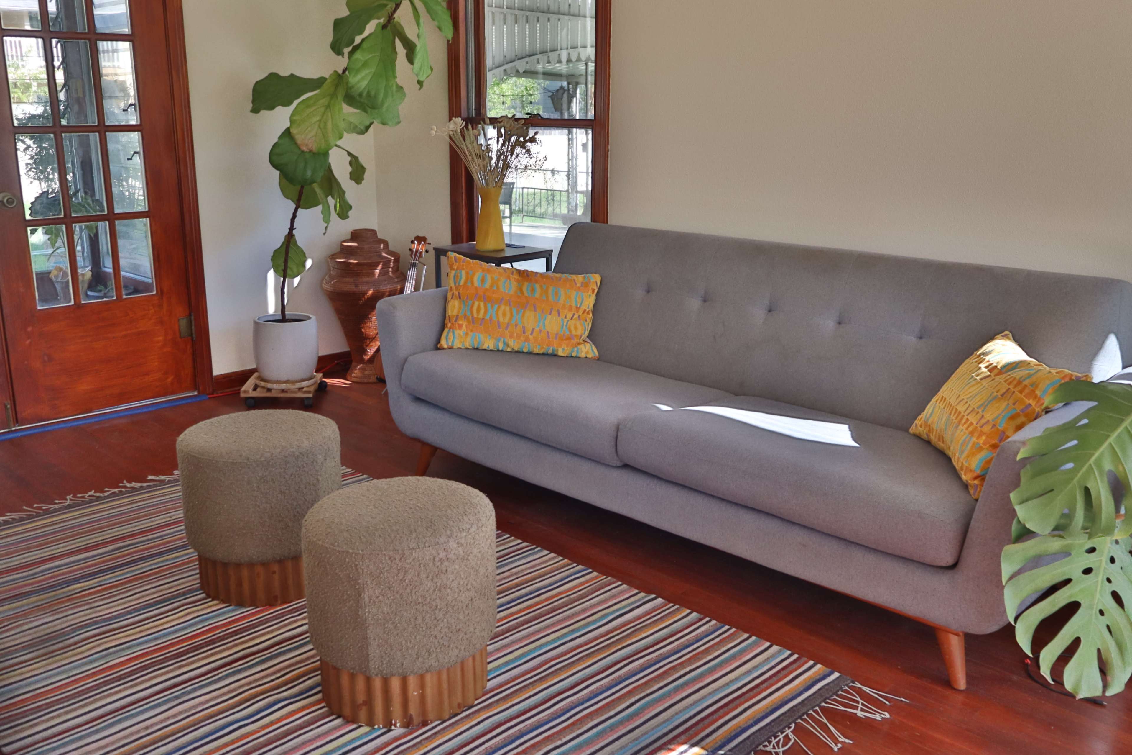 The image shows a cozy living room featuring a gray sofa with decorative pillows, two round stools, and a striped area rug, alongside plants and wooden doors.