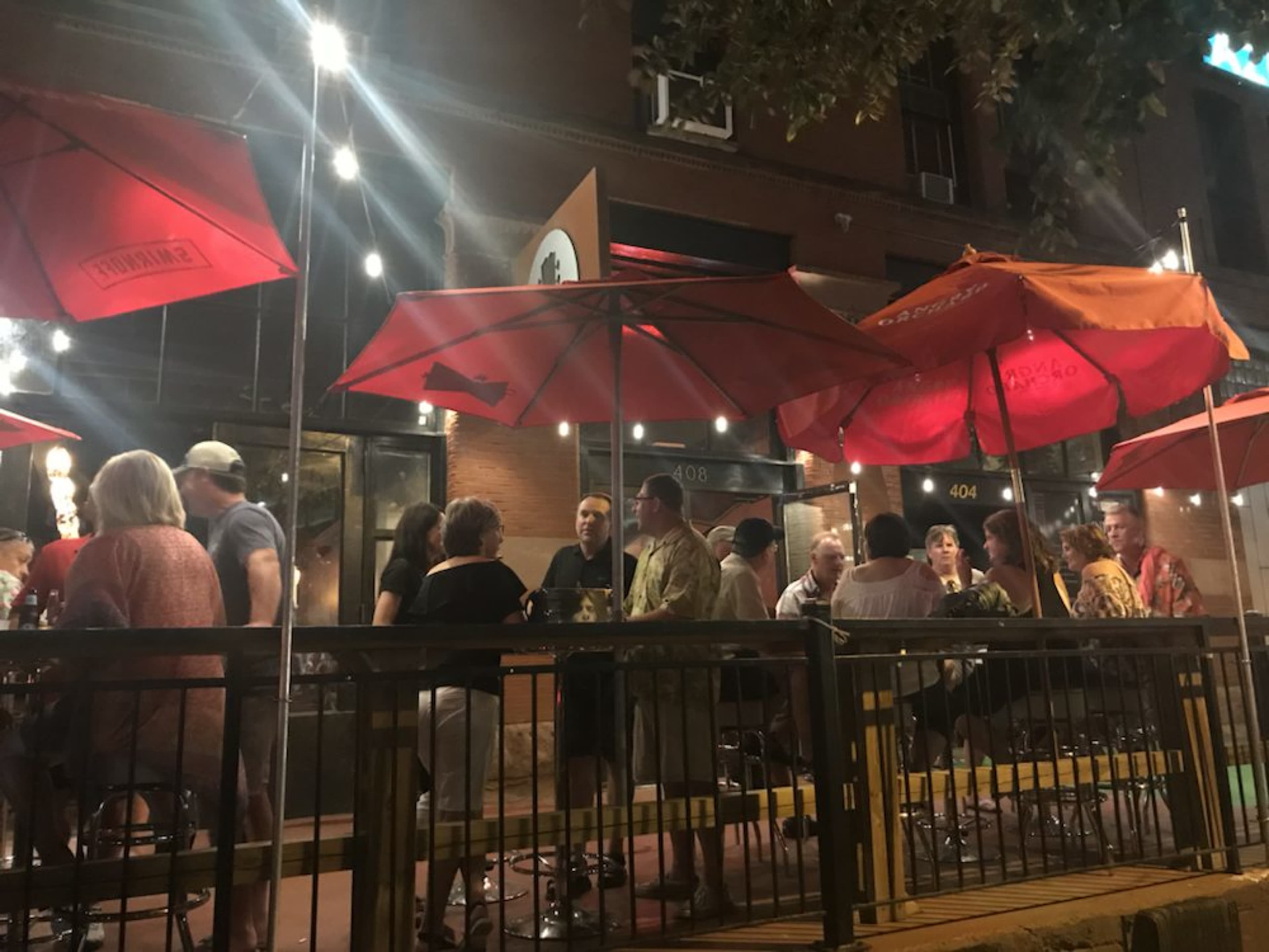 A group of people gathers under red umbrellas at an outdoor restaurant in the evening.