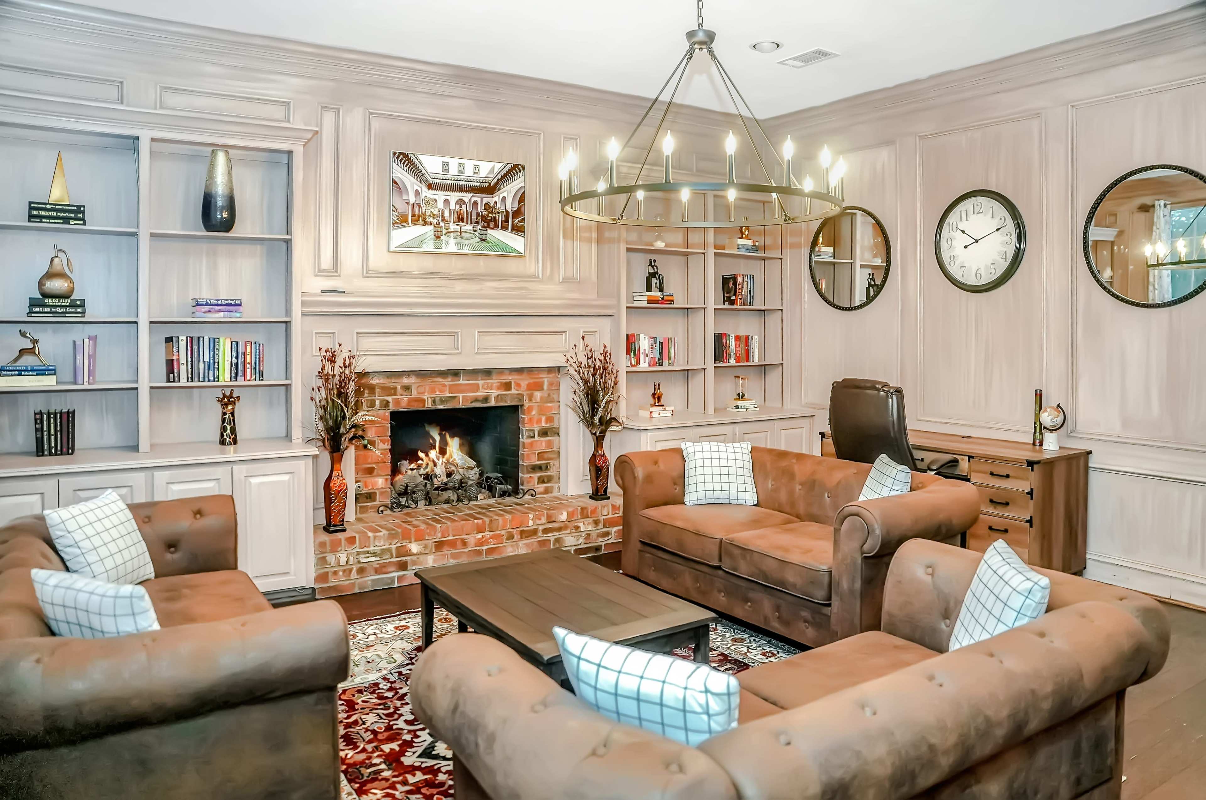 The image shows a cozy living room featuring a brick fireplace, two brown leather sofas, a wooden coffee table, and built-in bookshelves filled with various books and decor.