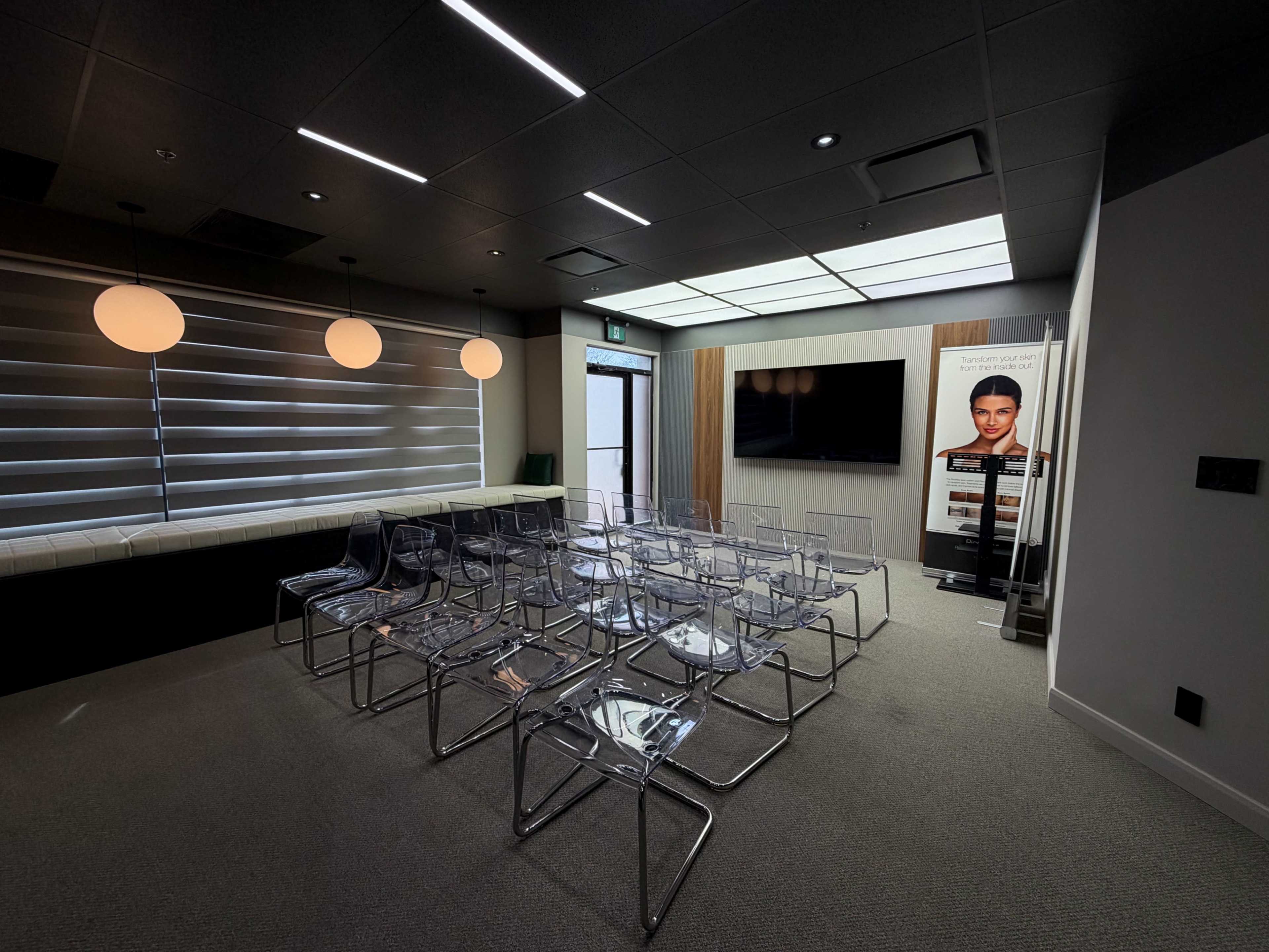 The image shows a modern meeting room with transparent chairs arranged around a central space, featuring a large wall-mounted screen and a poster on the side.