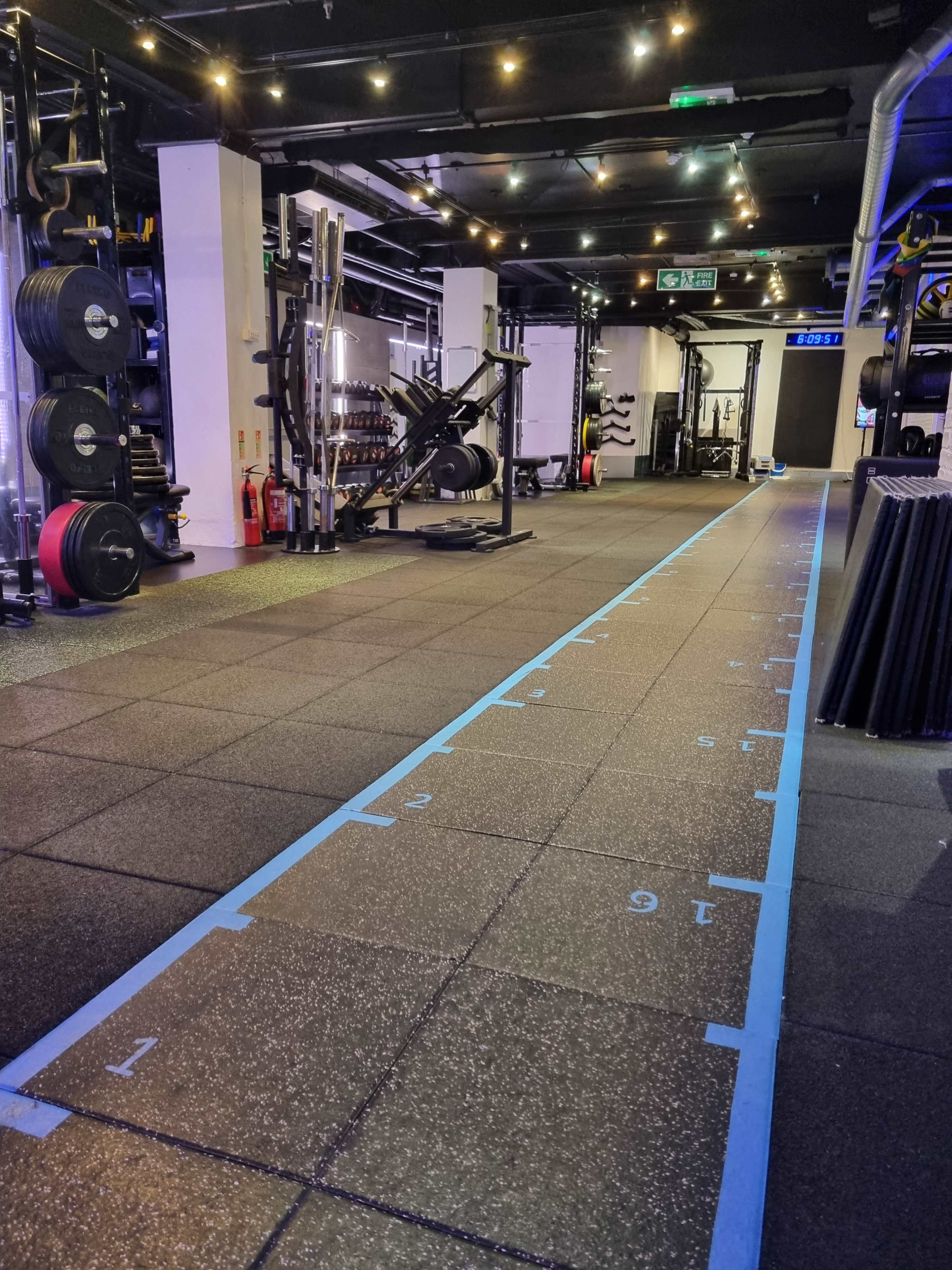 The image shows a gym floor with numbered blue markers on black rubber tiles and various weightlifting equipment arranged along the walls.