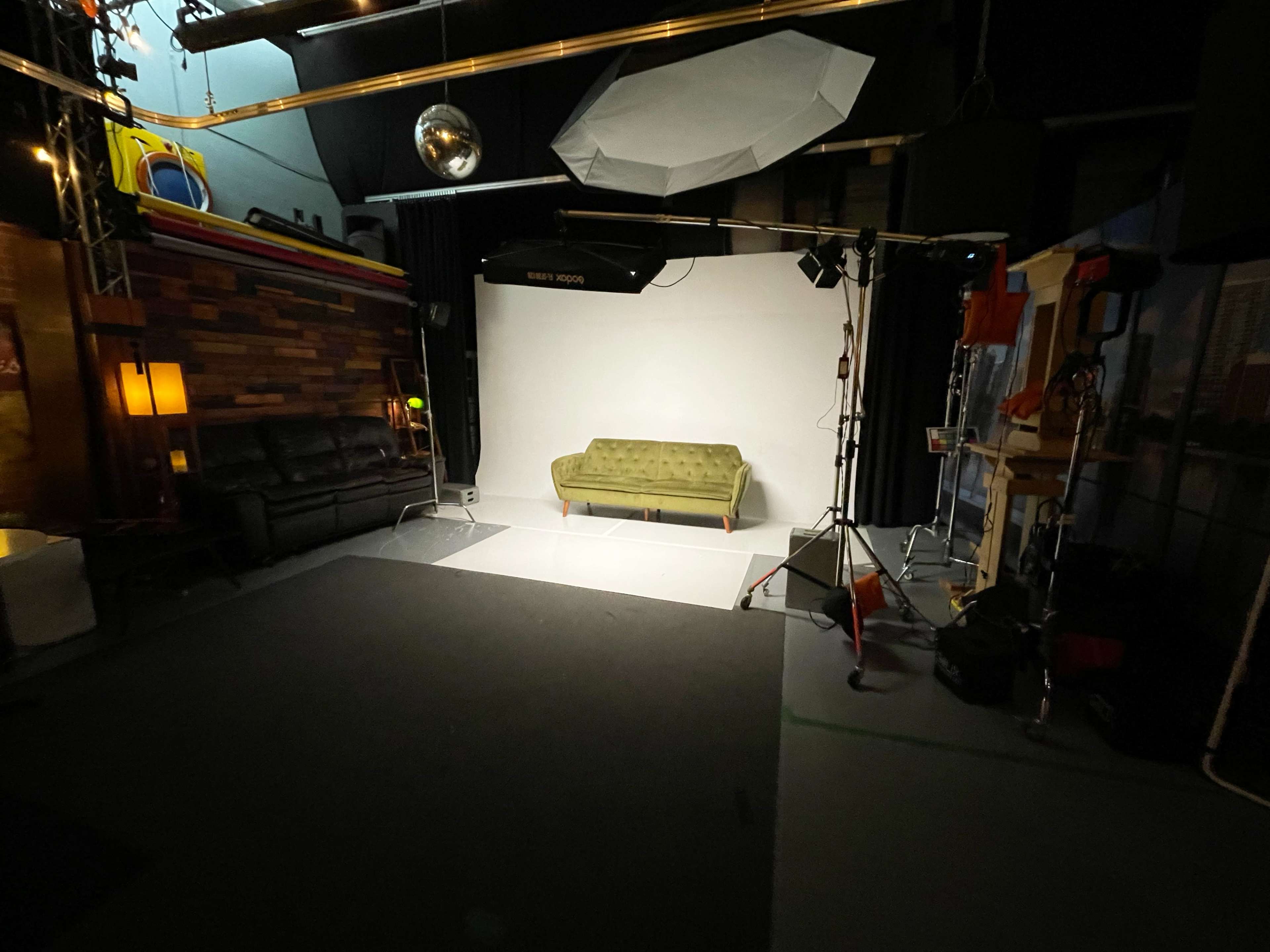 The scene features a dimly lit studio space with a green couch positioned in front of a white backdrop, surrounded by various lighting equipment and props.