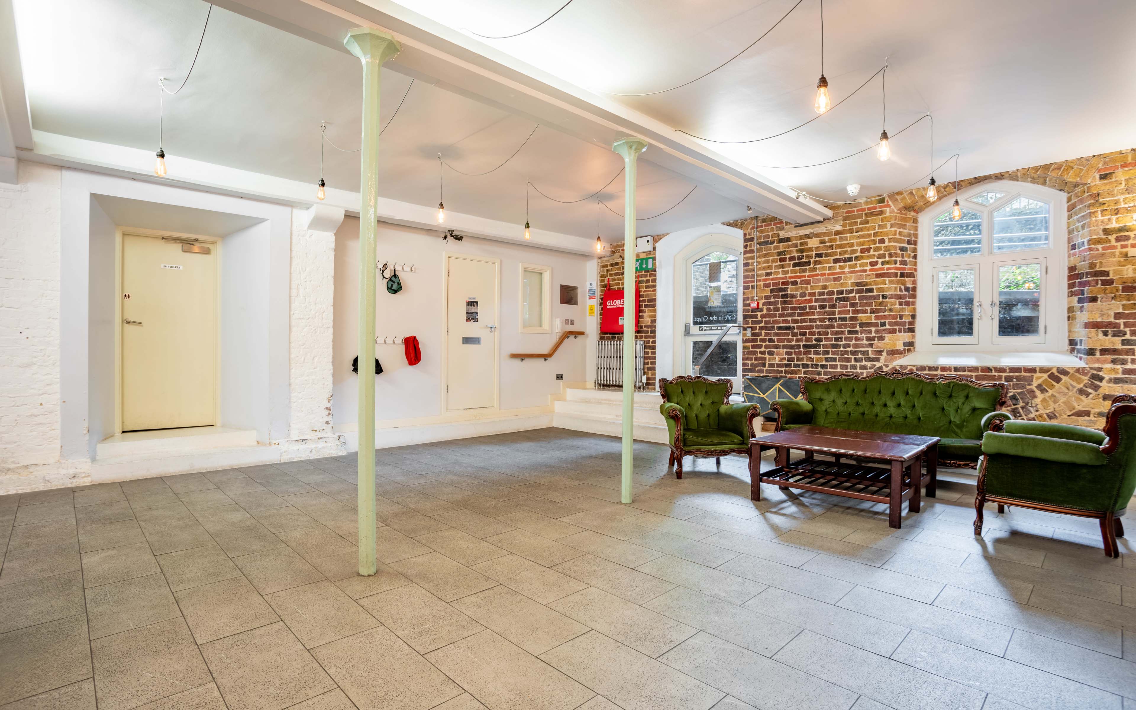 A spacious interior with stone flooring, a green velvet sofa set, and exposed brick walls, illuminated by hanging light bulbs.