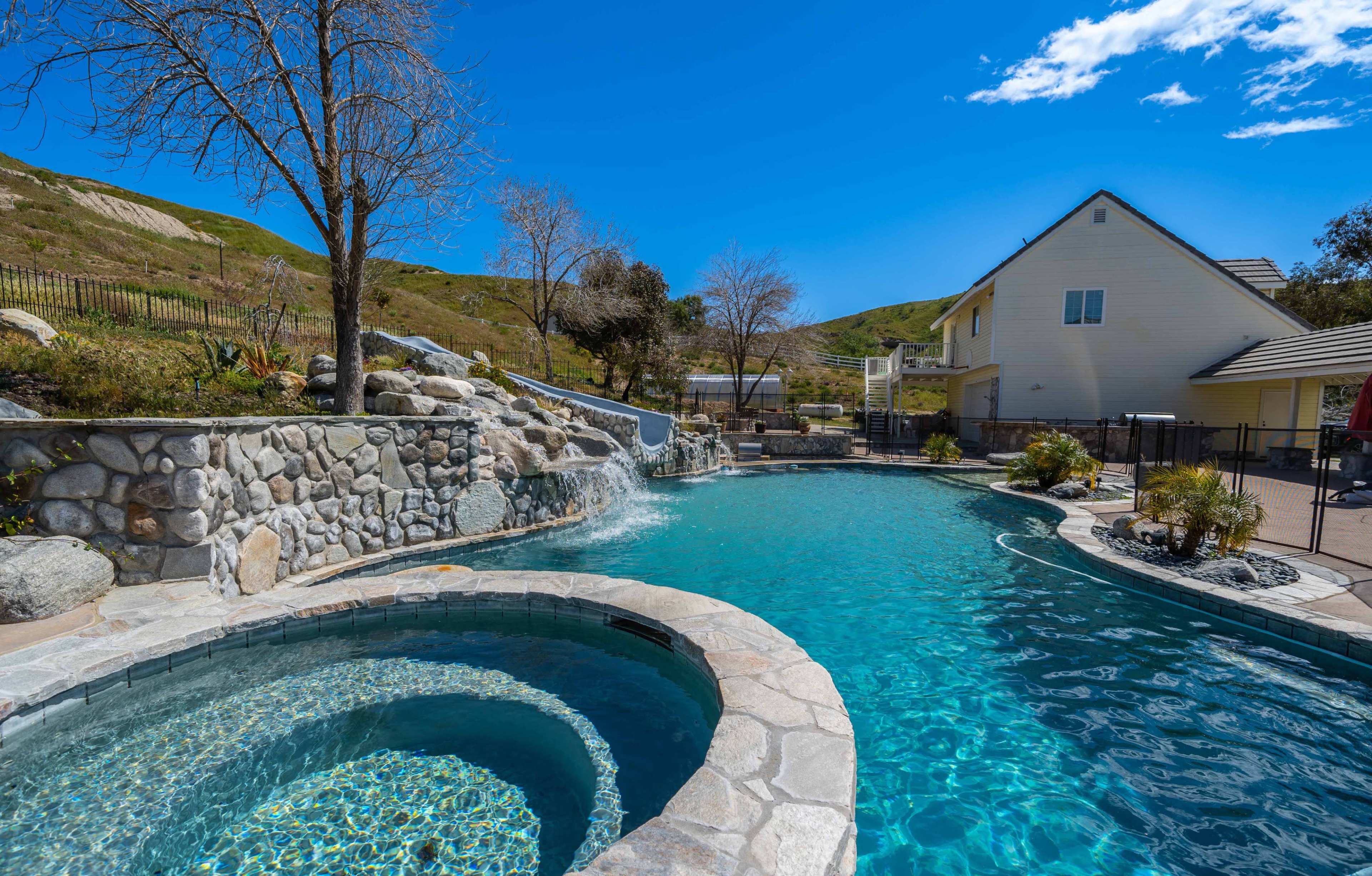 The image shows a large backyard pool with a spa, surrounded by stone features and a hillside in the background.