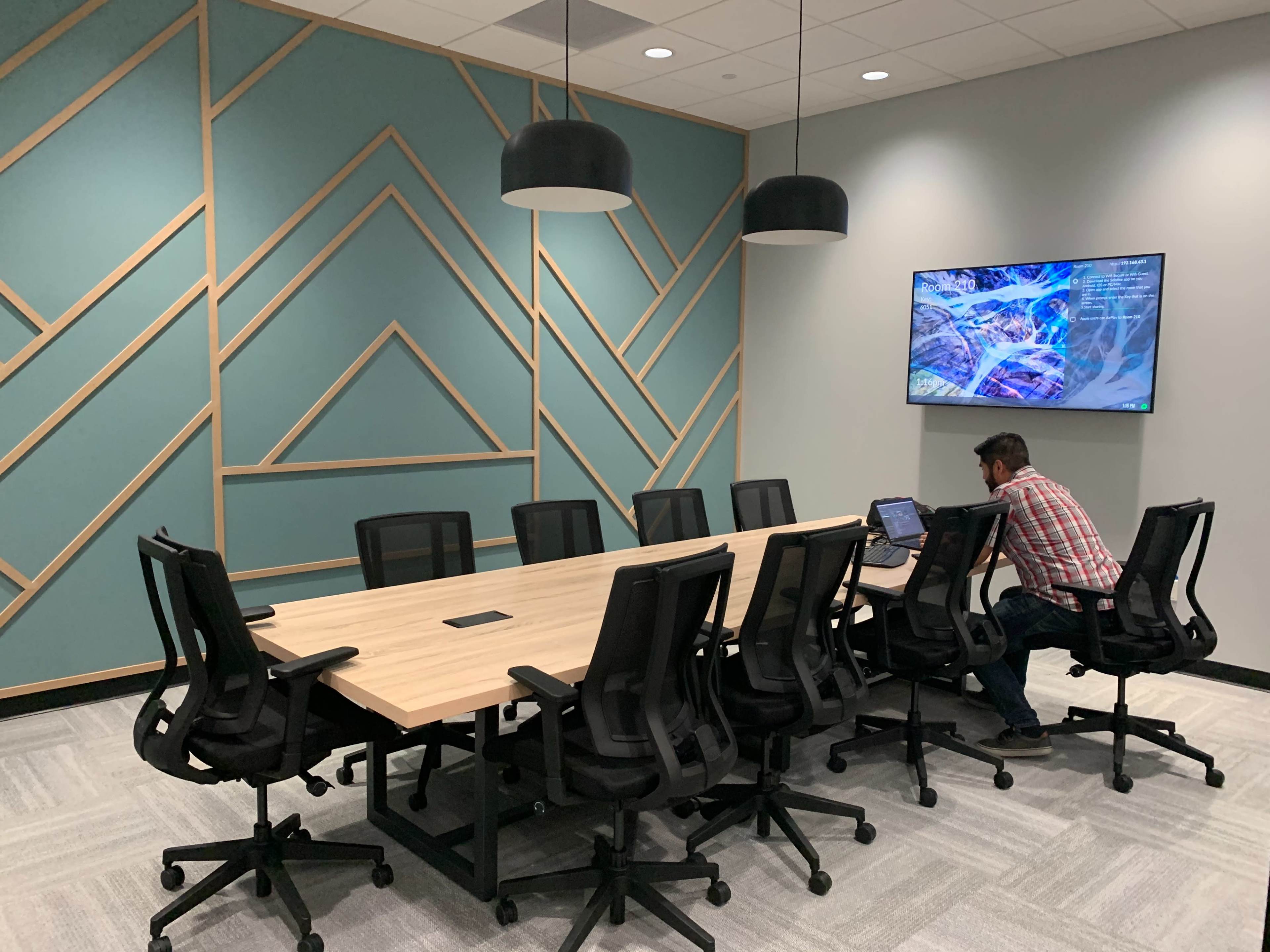A conference room features a long wooden table surrounded by black office chairs, with a large screen mounted on the wall displaying digital content.