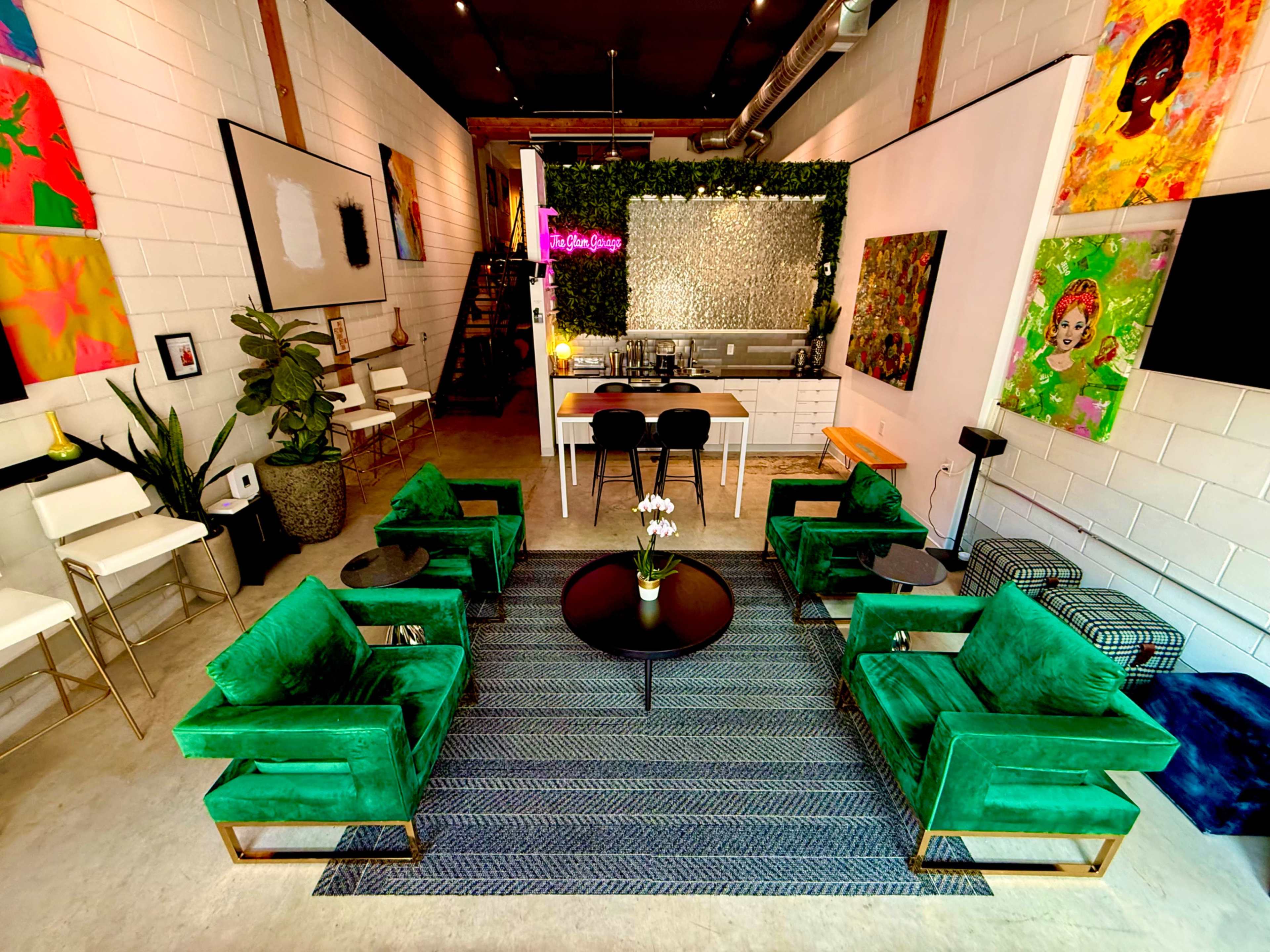 The image shows a modern, vibrant interior space with green velvet chairs arranged around a circular table, a kitchen area in the background, and colorful artwork on the walls.