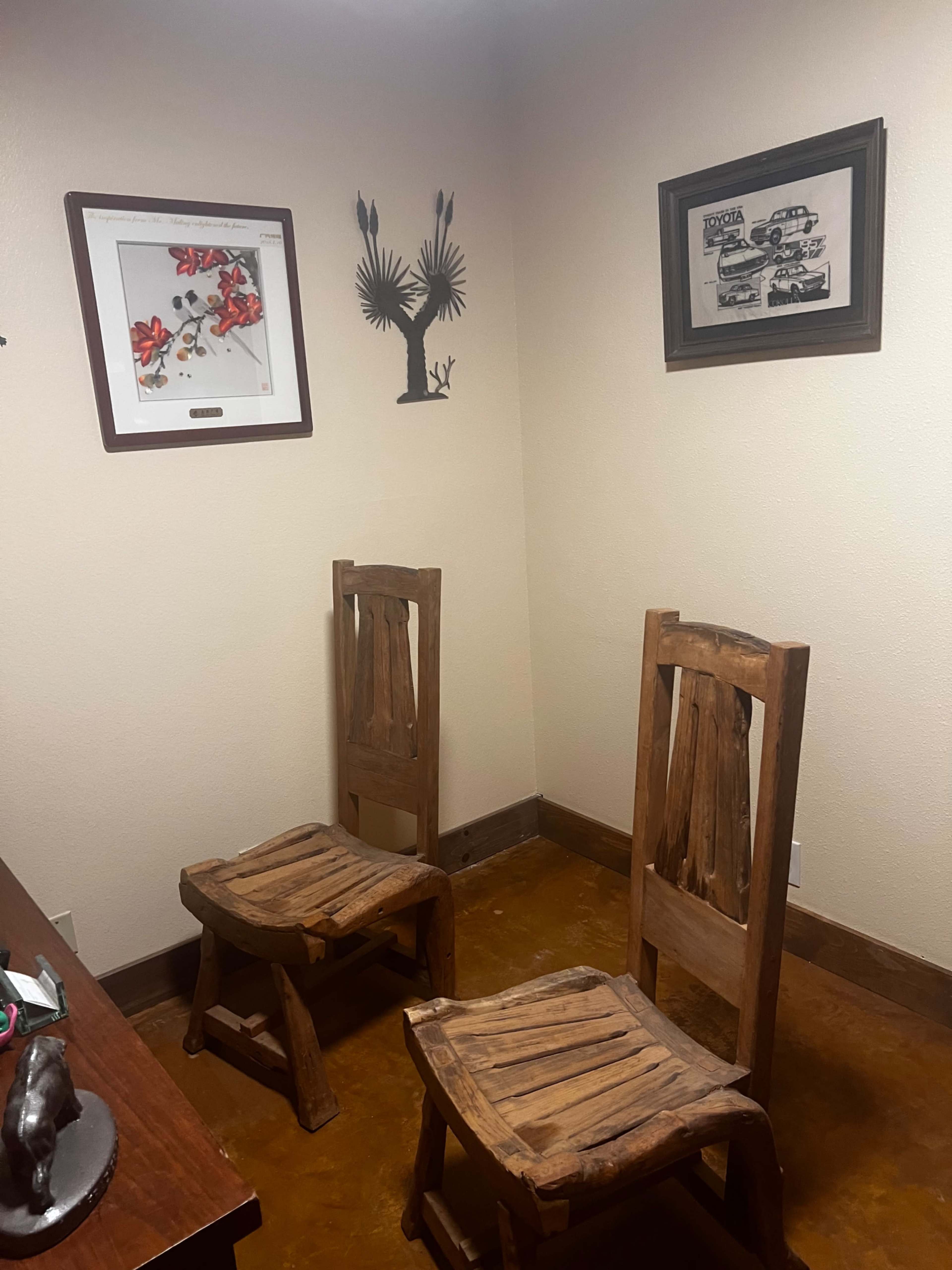 The image shows a small, empty room with two wooden chairs facing each other, decorated with framed artwork on the walls.