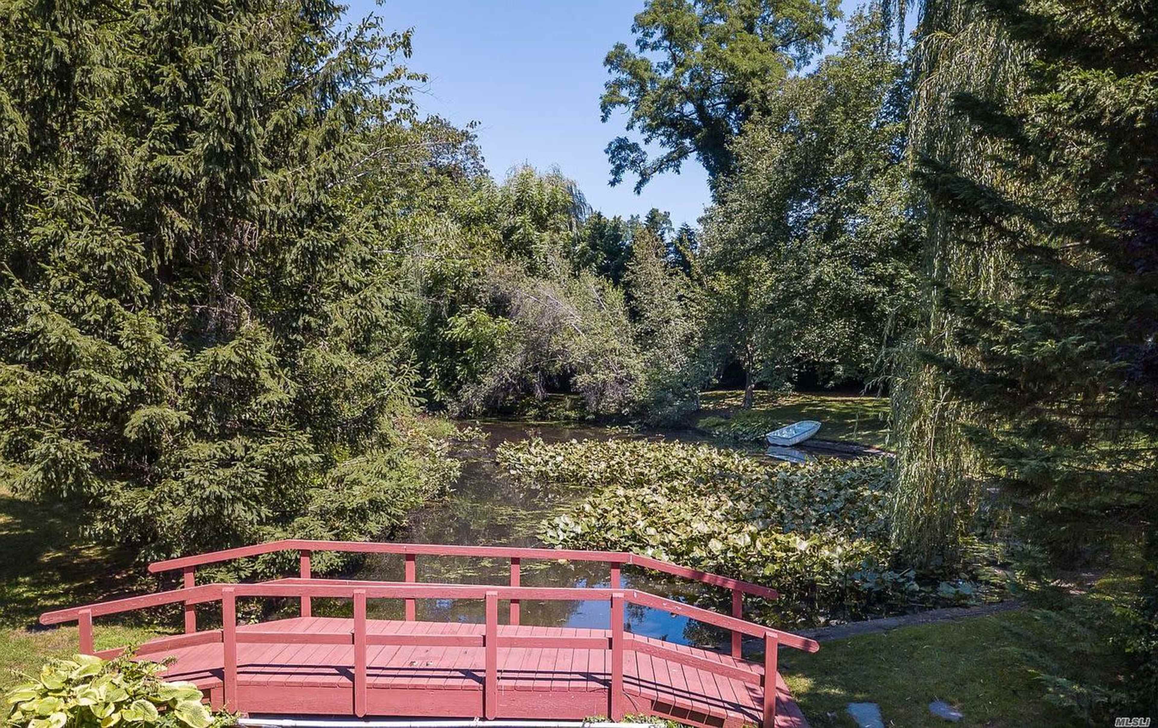 A red wooden bridge spans a small pond surrounded by lush greenery and a white boat rests at the water's edge.