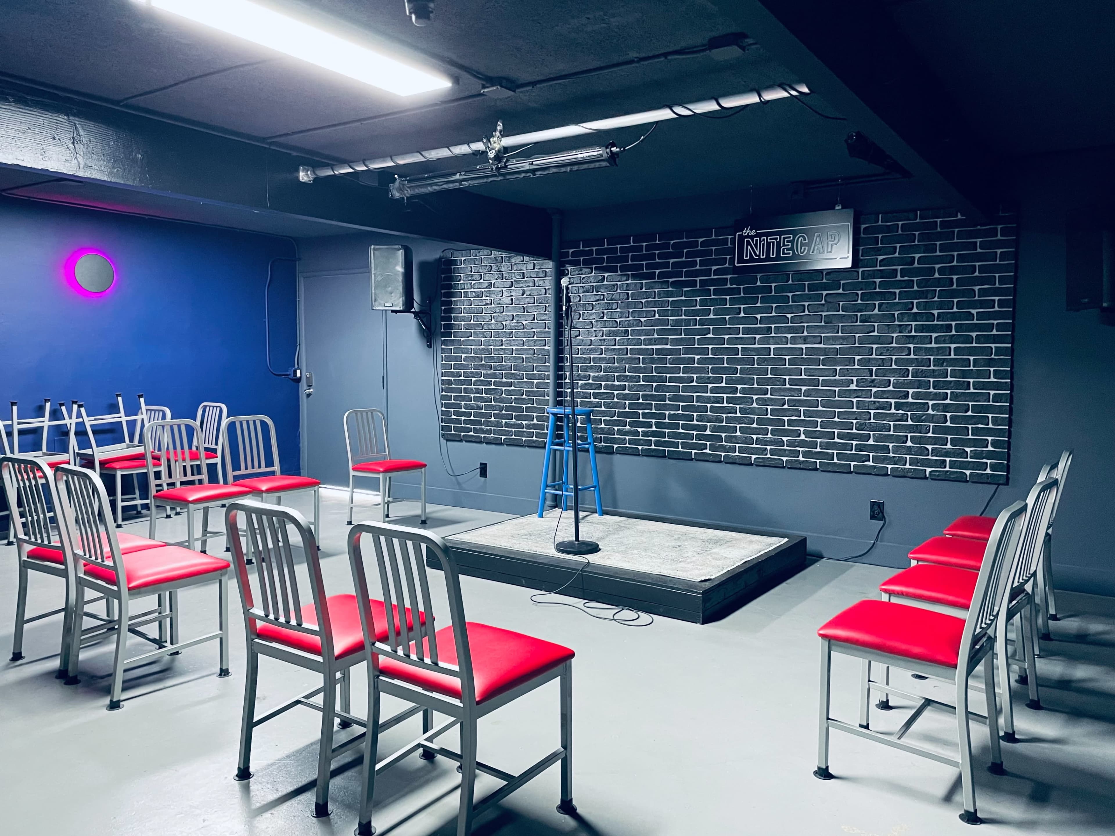 The image shows a small comedy club with rows of red-seated chairs facing a microphone on a stage against a brick wall.