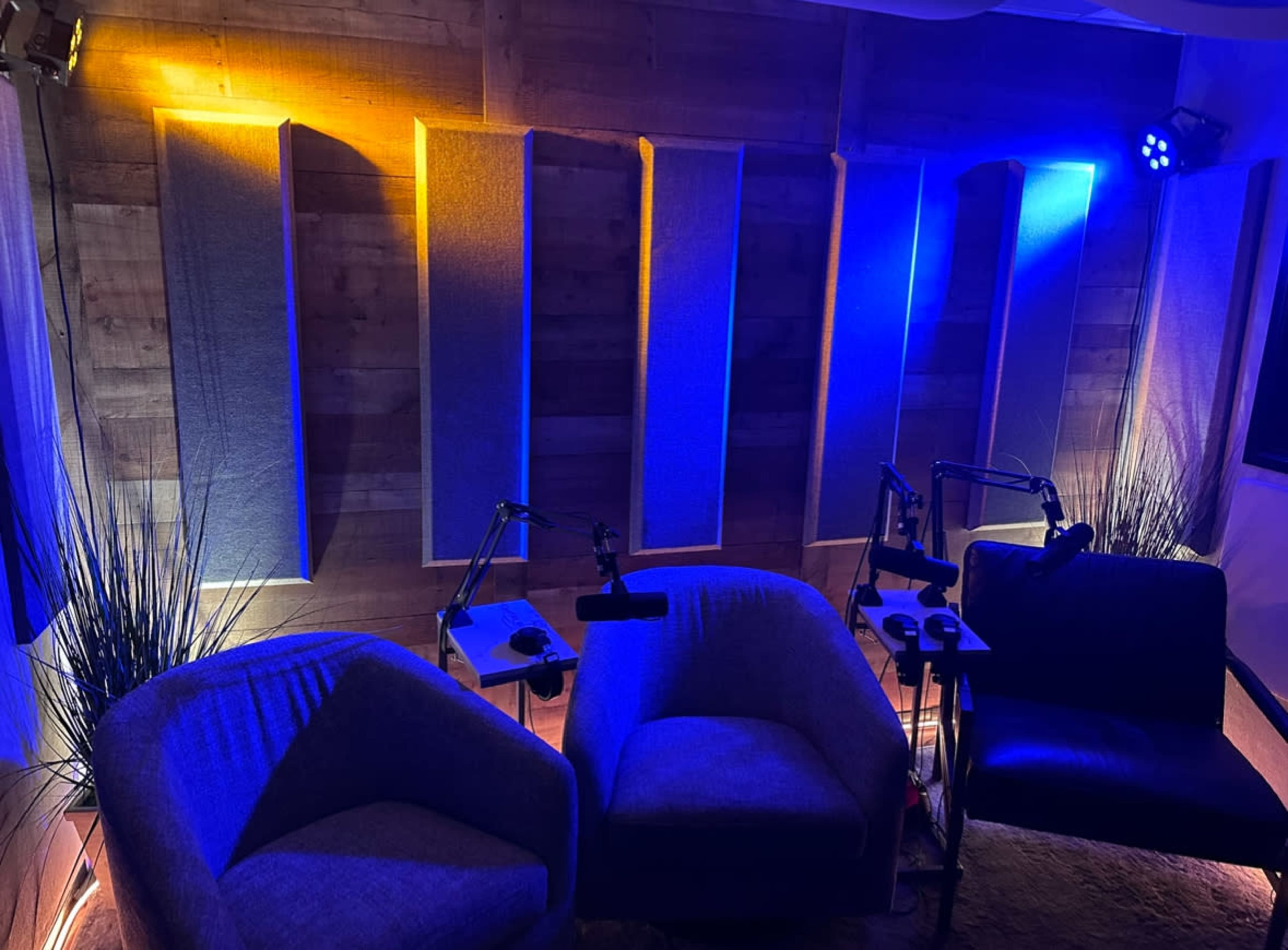 The image shows a cozy recording studio with two blue armchairs and soundproof wall panels illuminated by warm yellow and cool blue lights.